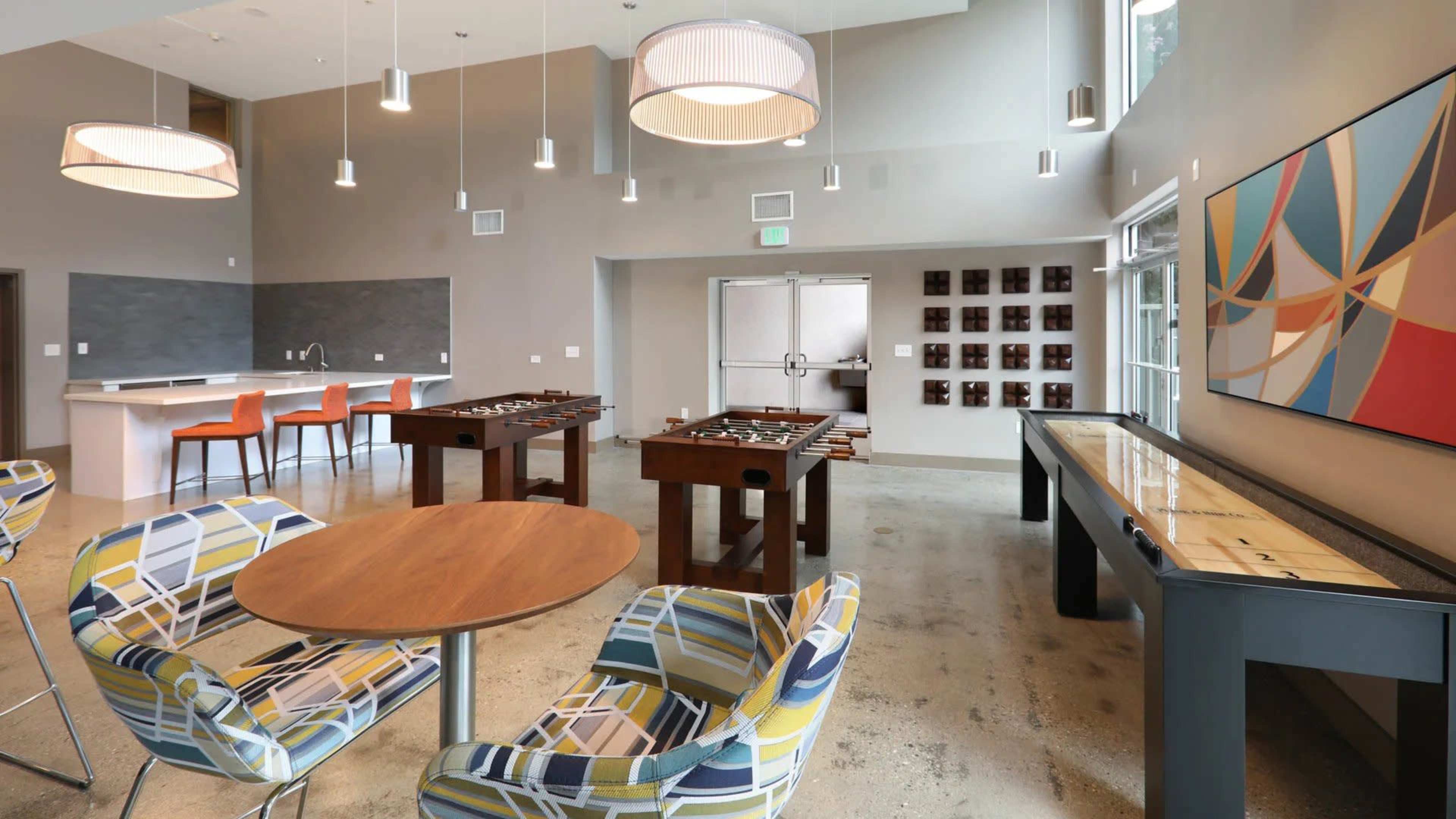 The image shows a modern recreational room with a dining table, colorful chairs, foosball tables, and a shuffleboard table, all set in a well-lit space.