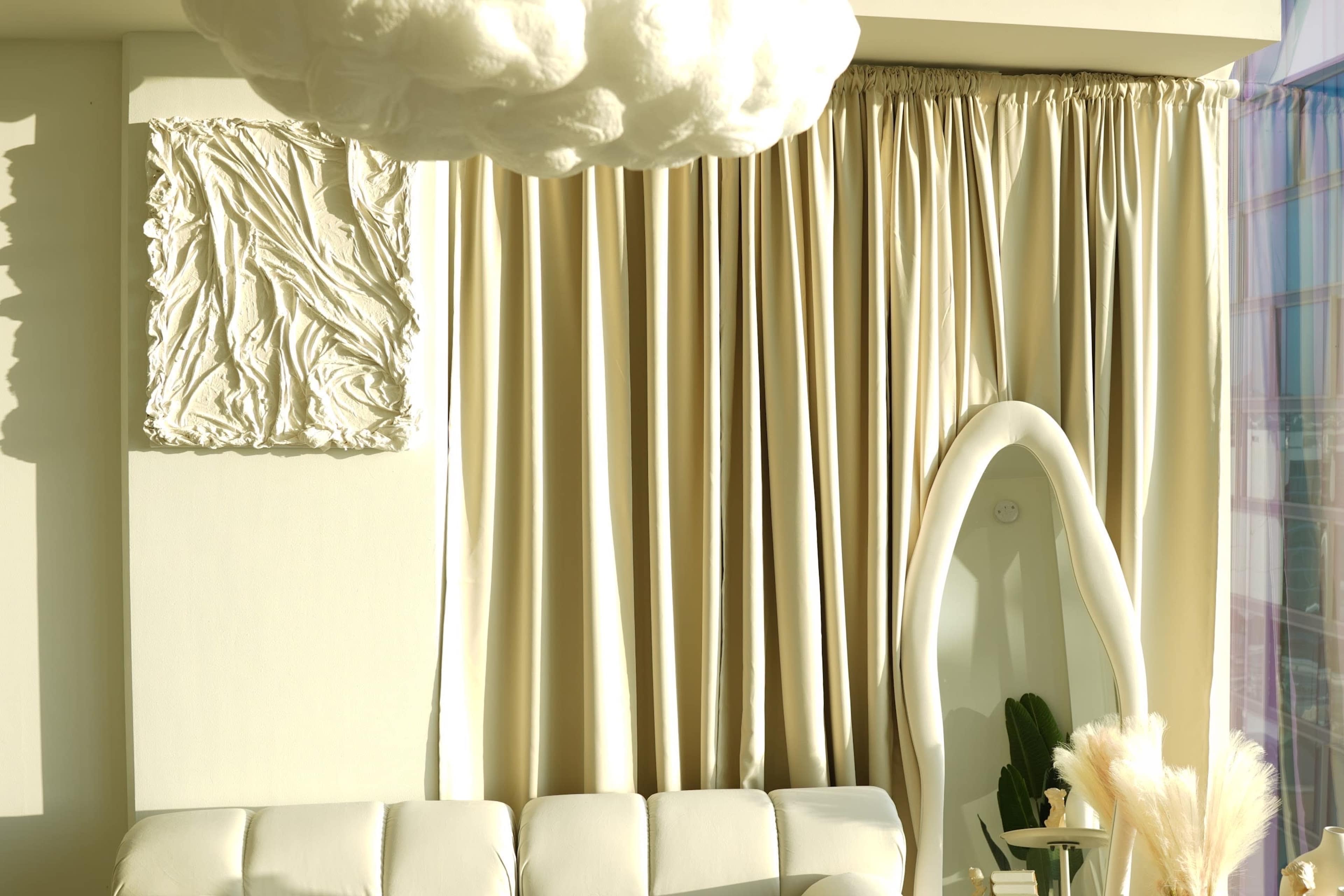The image features a bright interior with a white couch, cream-colored curtains, a textured wall art piece, and a unique, pod-like chair.
