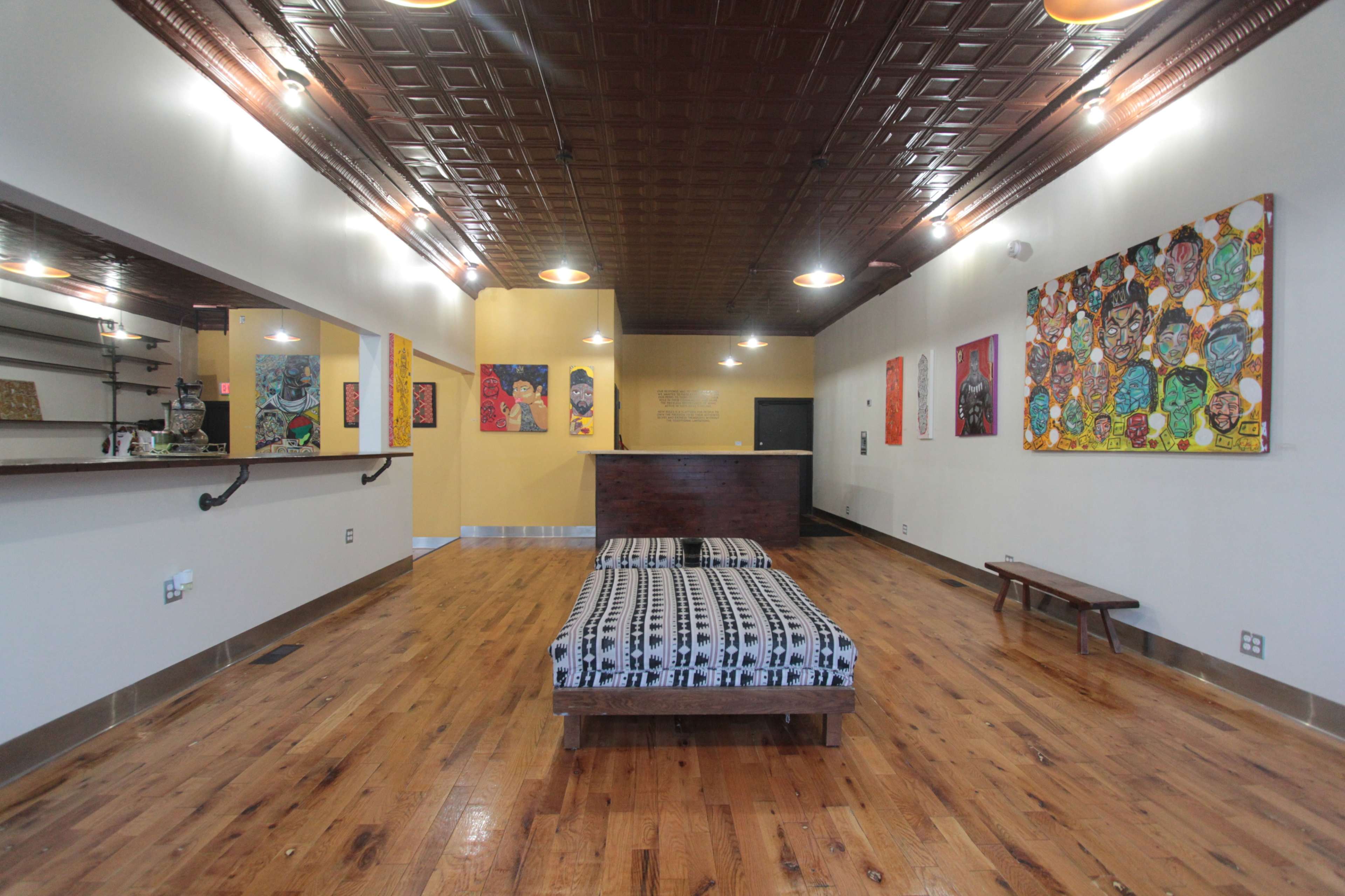 The image shows a spacious interior of a room with wooden floors, a patterned mattress on a wooden platform, a reception area on the left, and colorful artwork displayed on the walls.