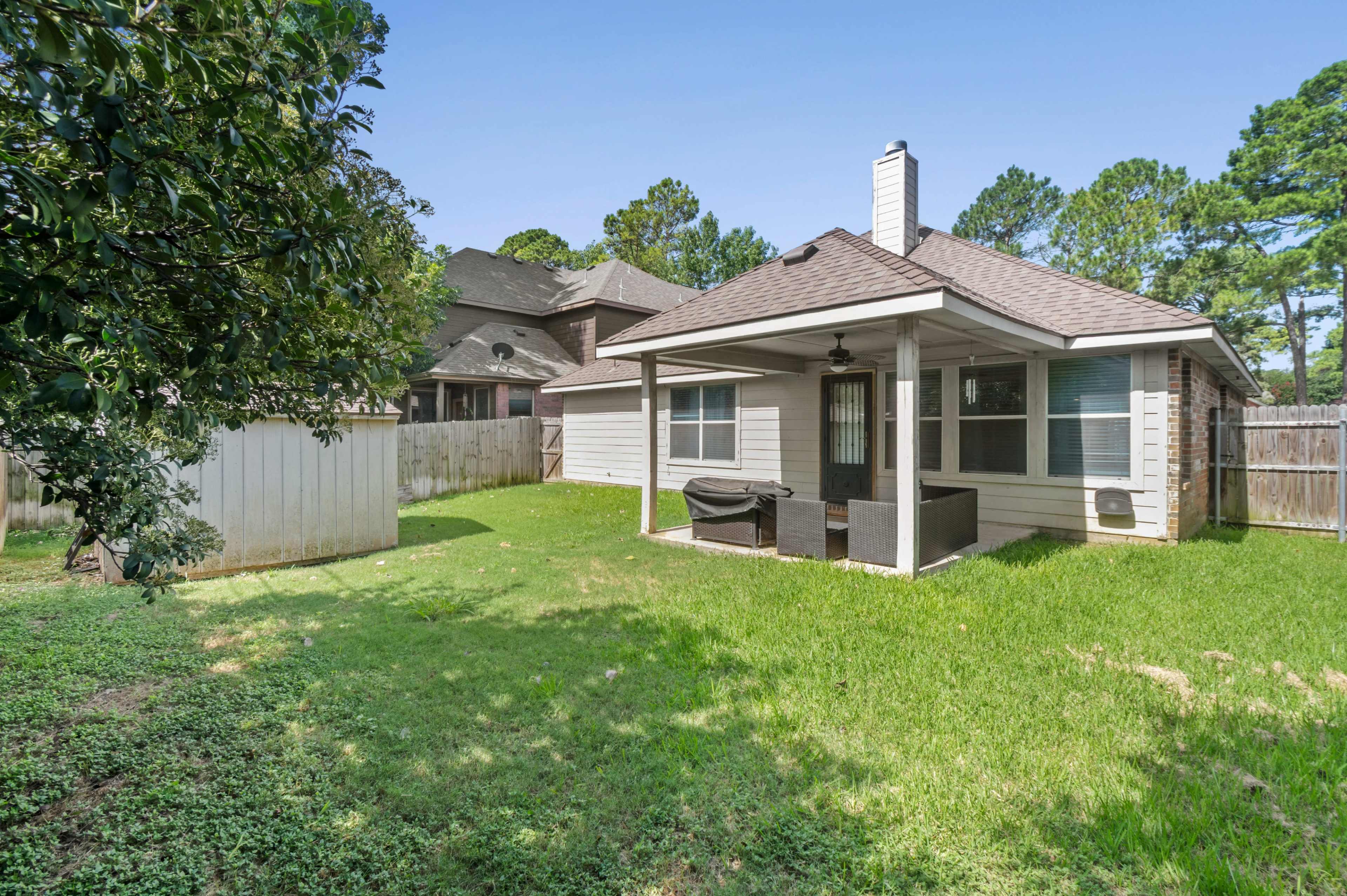 The image shows a backyard with a patio area covered by a roof, surrounded by green grass and a wooden fence.