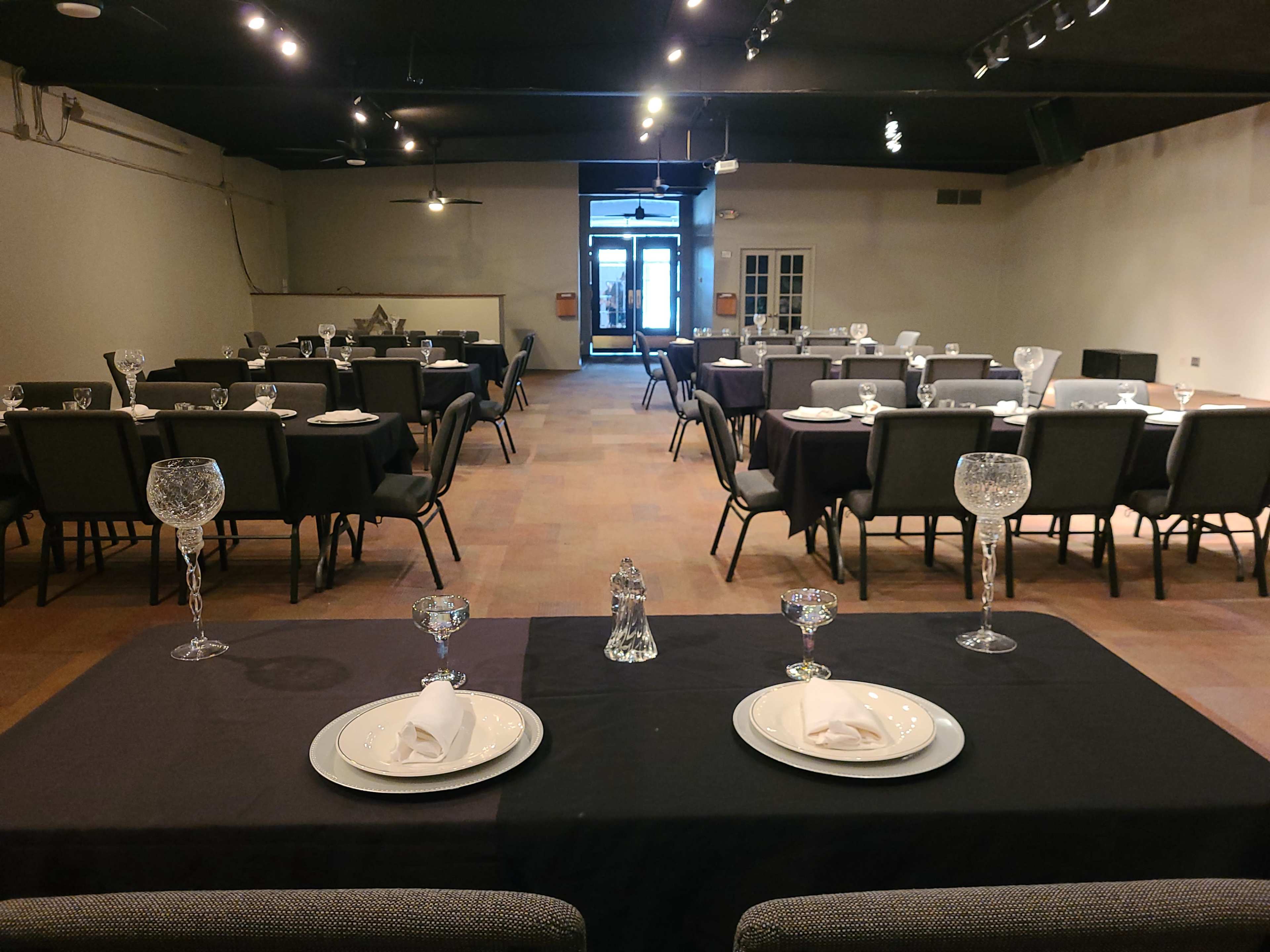 The image shows a banquet room set with tables arranged for dining, featuring white plates, crystal glassware, and neatly folded napkins.