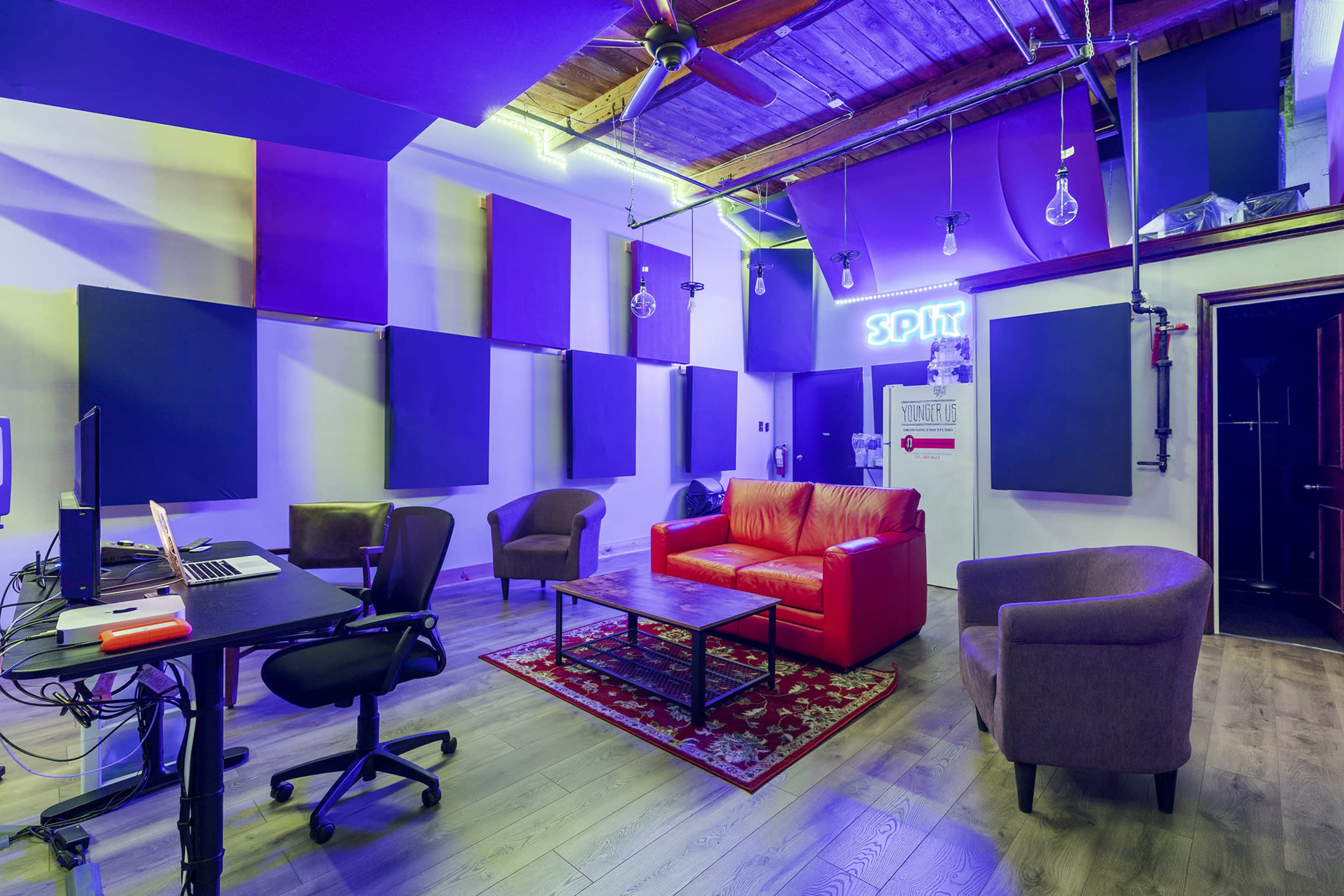 A recording studio features a red couch, two armchairs, a coffee table, and various soundproofing panels on the walls, with neon lighting and exposed bulbs overhead.