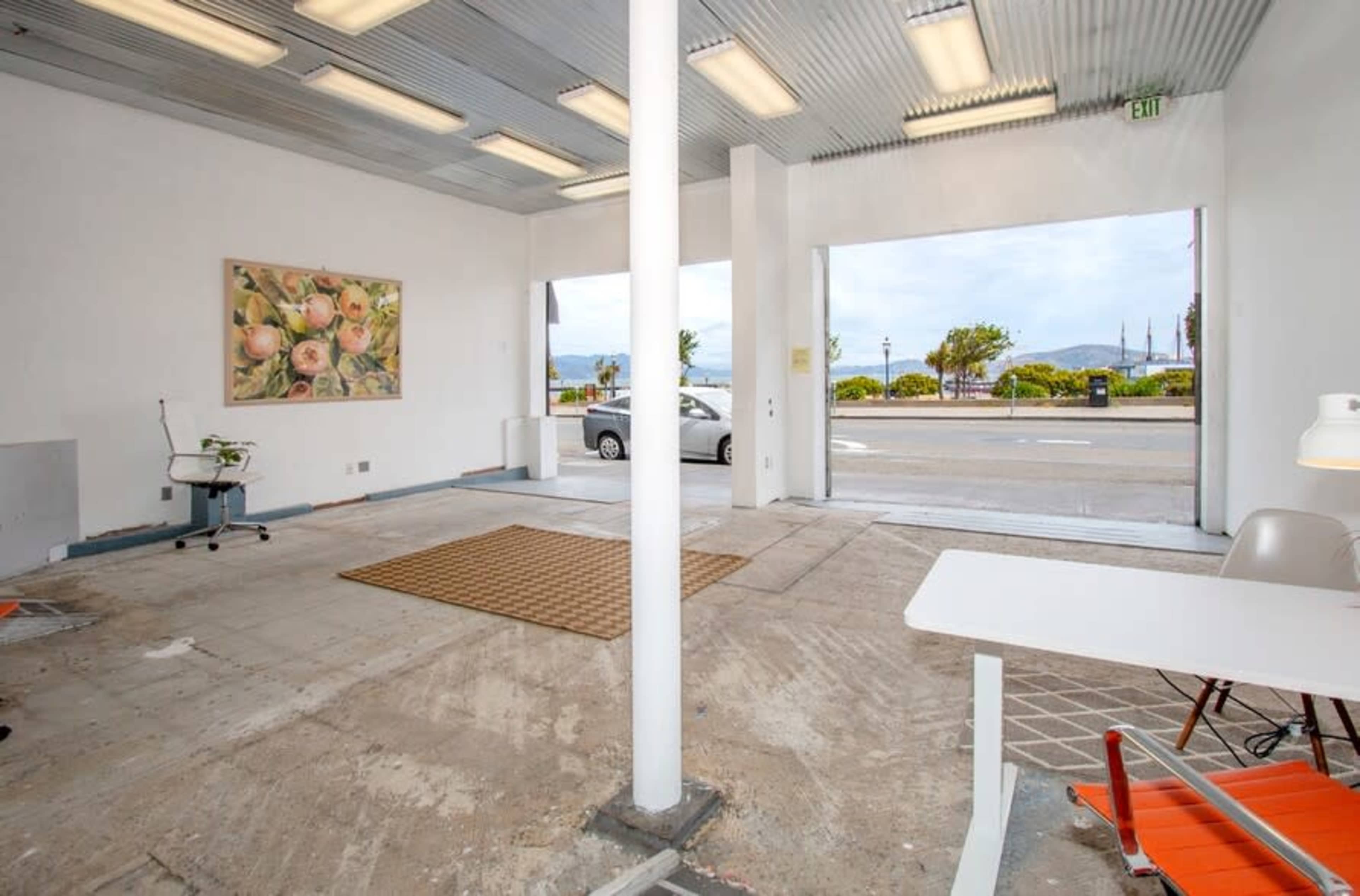 The image shows an empty room with a concrete floor and large openings leading to a view of an outdoor area with vehicles and trees.