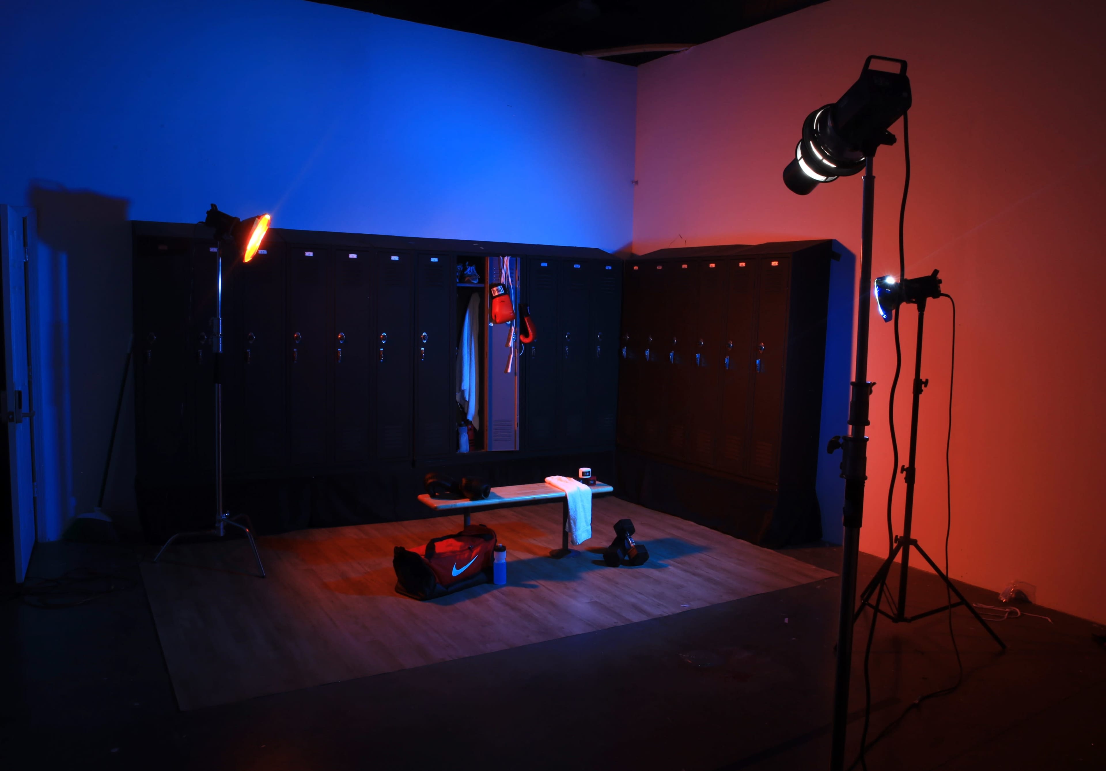 A fitness locker room scene features lockers lined against a wall, illuminated by colored stage lights, with workout equipment and a towel on a bench in the center.