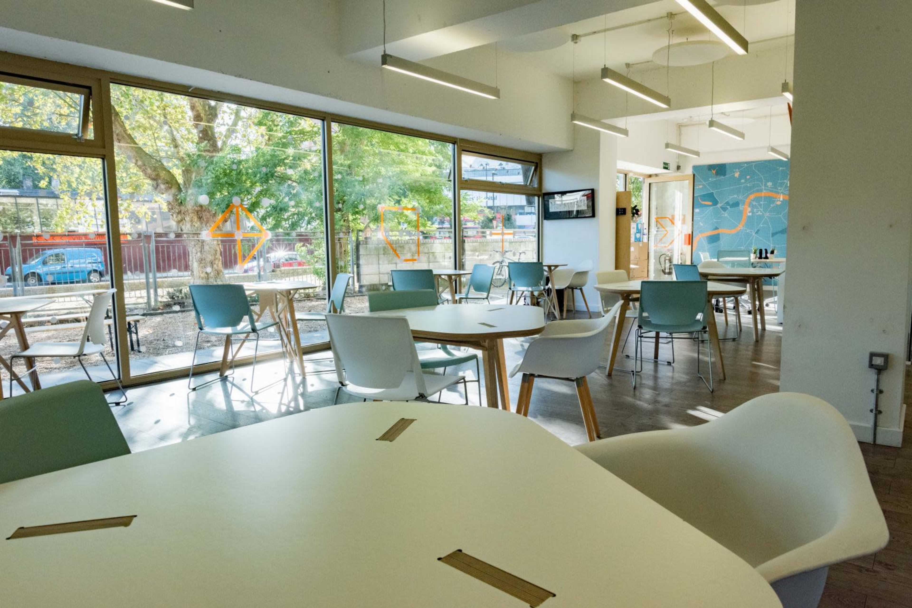 The image shows a bright, modern café interior with several tables and chairs arranged around large windows overlooking a tree-lined area.