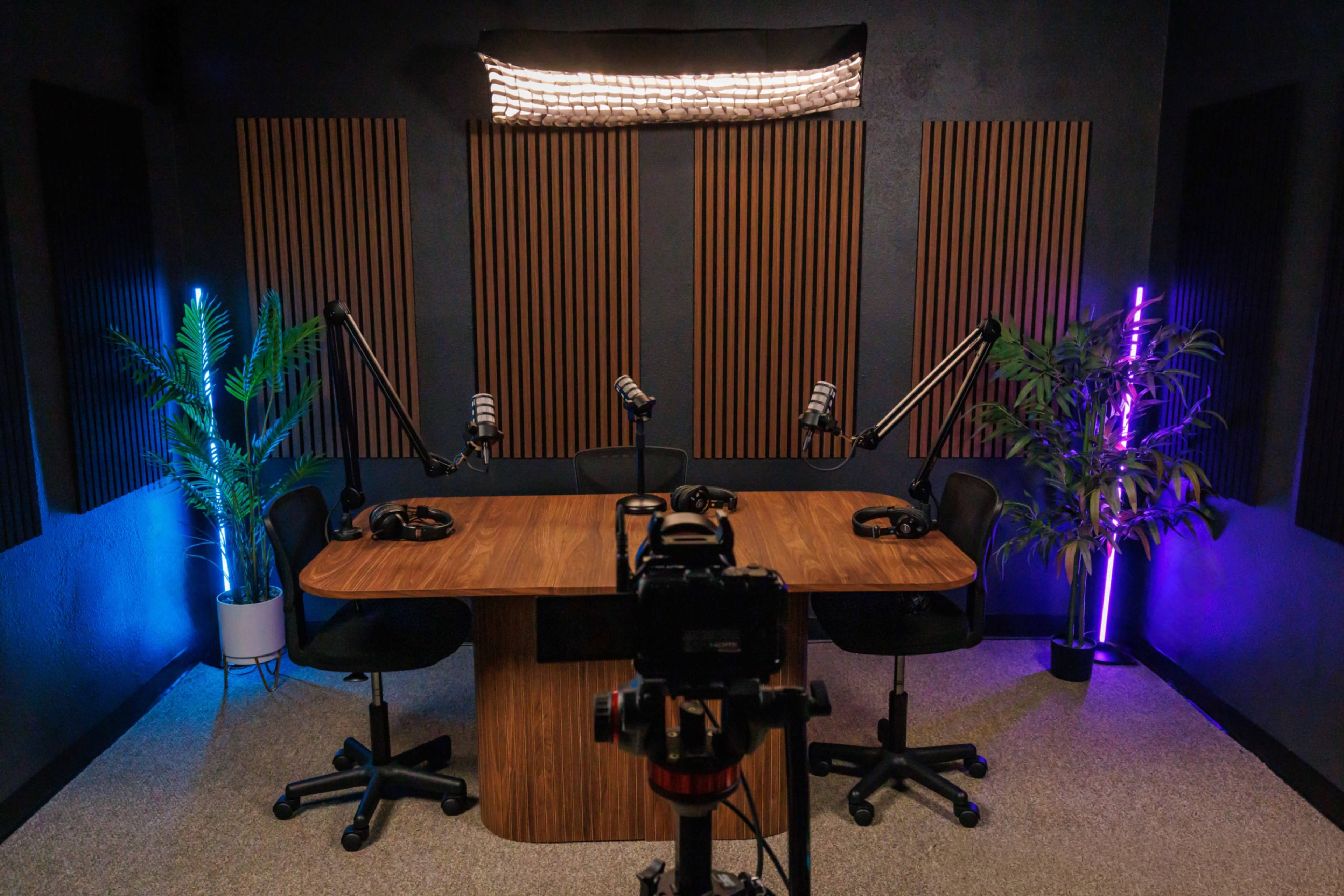 The image shows a podcast studio with a wooden table, two black chairs, and microphone stands, complemented by colorful lighting and soundproofing panels on the walls.