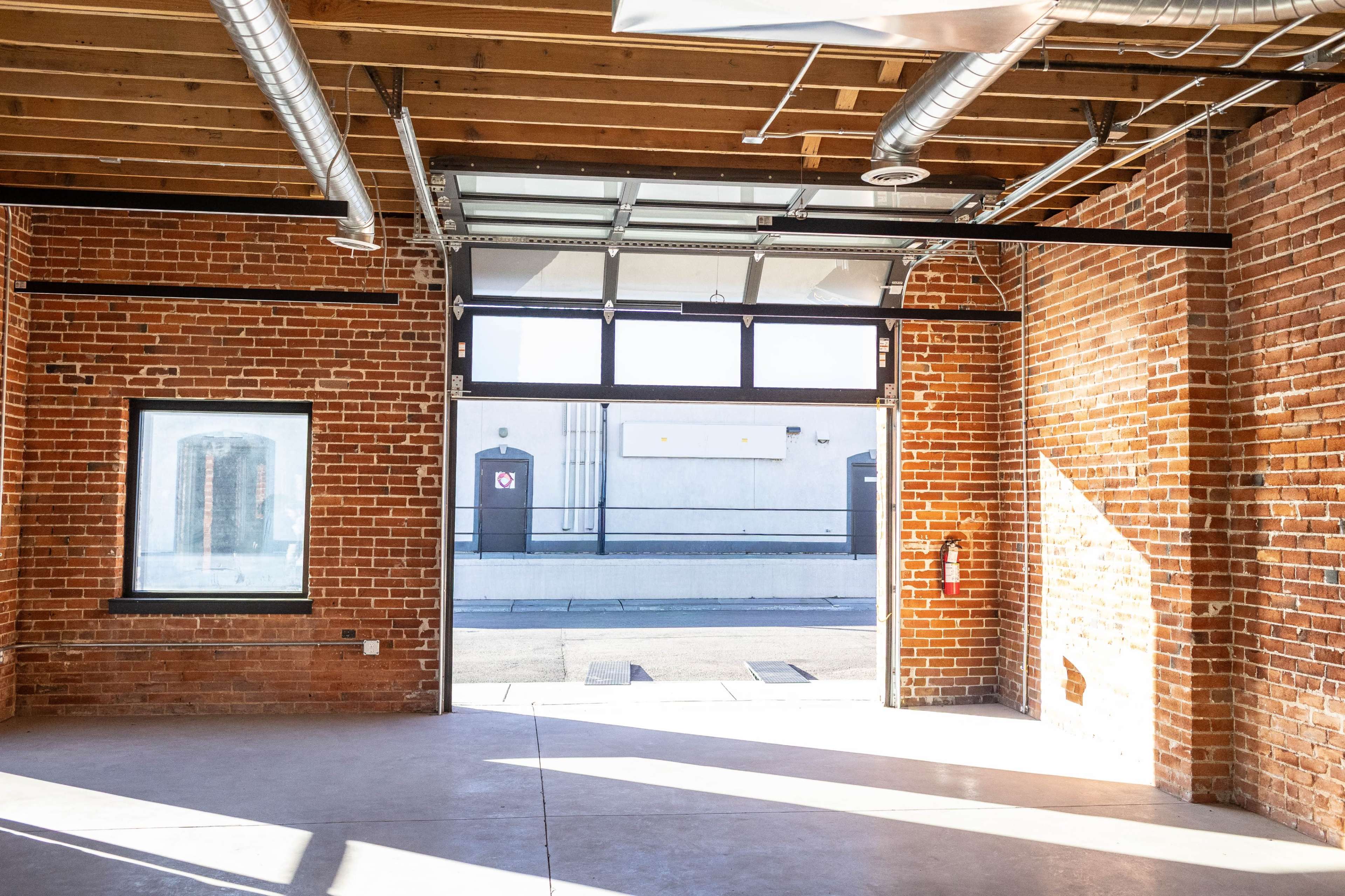 The image shows an industrial interior with exposed brick walls, large windows, and an open garage door revealing an exterior loading area.