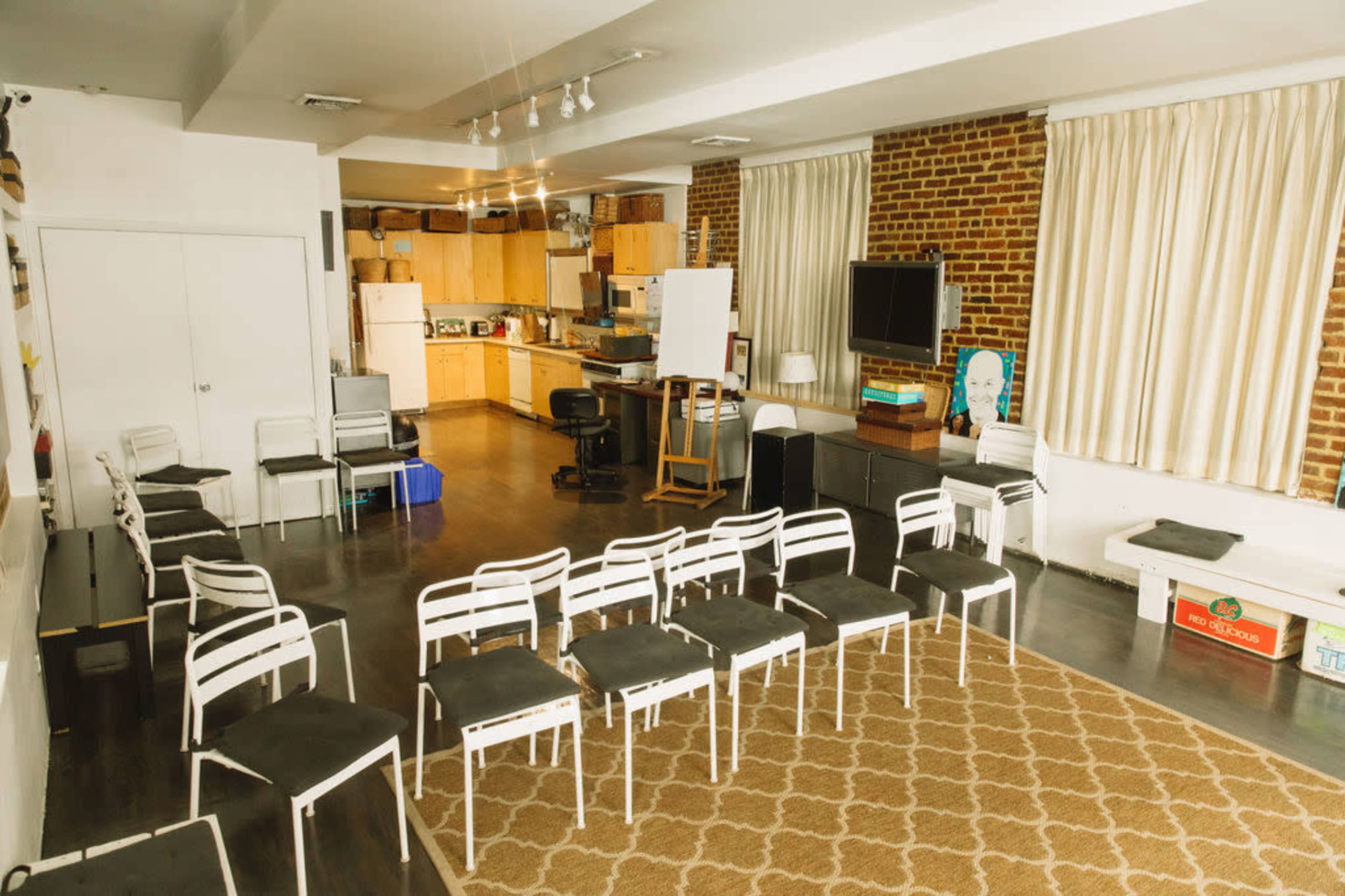 The image shows a spacious room with a kitchen area, a seating arrangement of white chairs facing a central space, and exposed brick walls.