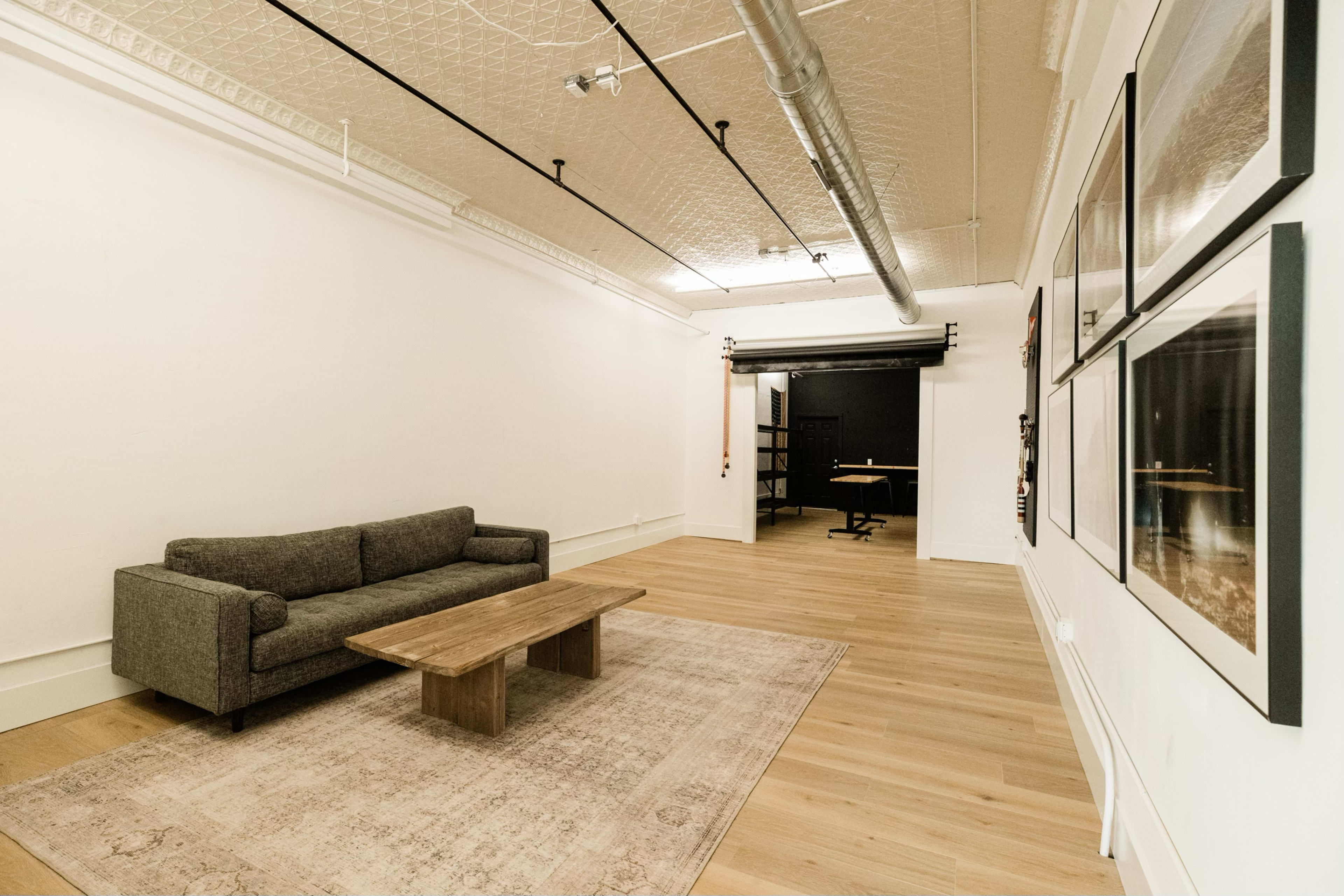 The image shows a spacious, minimally furnished room with a gray sofa, a wooden coffee table, and framed pictures on the walls, leading to a darker area at the back.