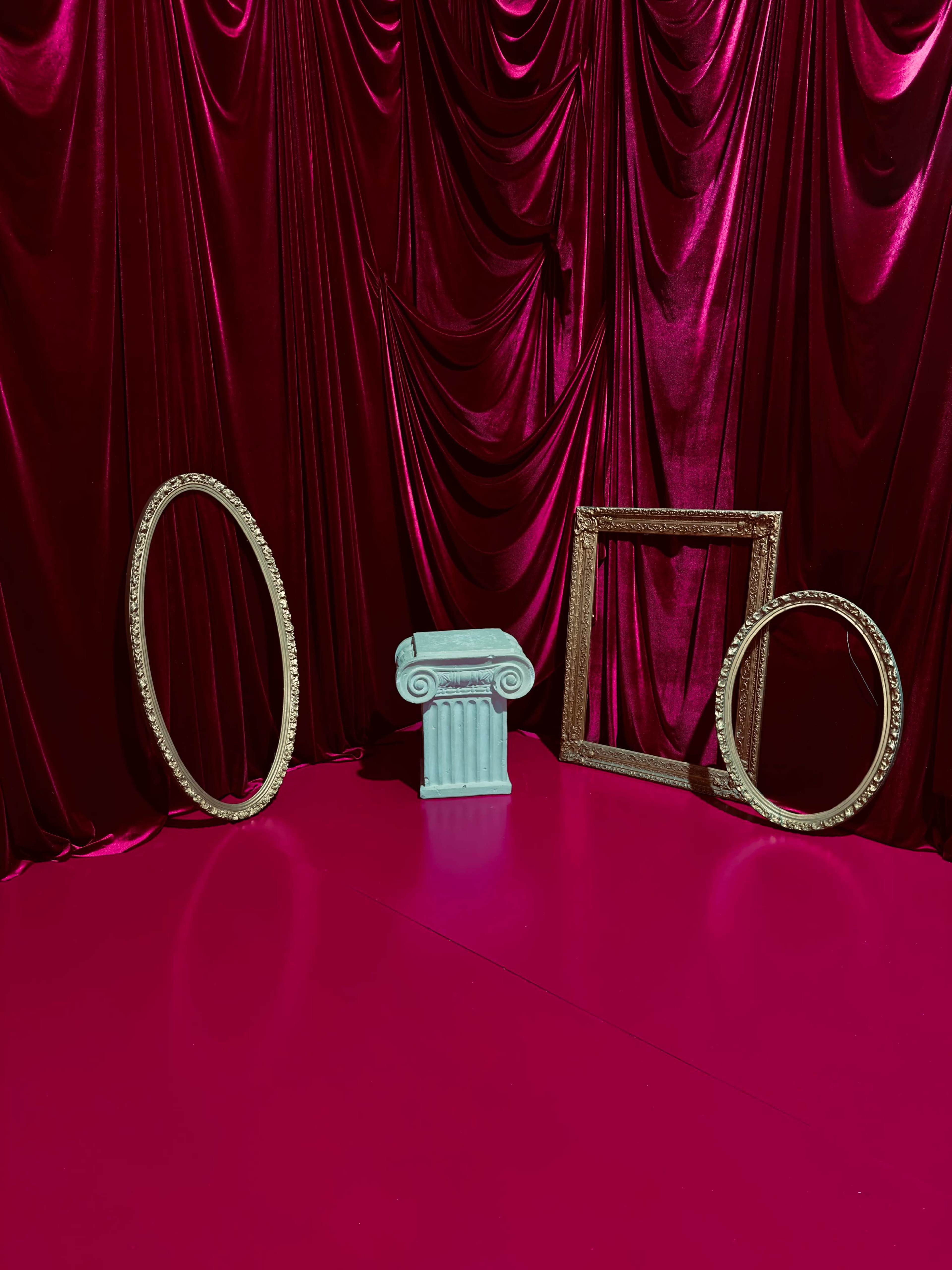 The scene features a pink floor with a central turquoise column and two ornate frames, set against a backdrop of deep red velvet drapery.