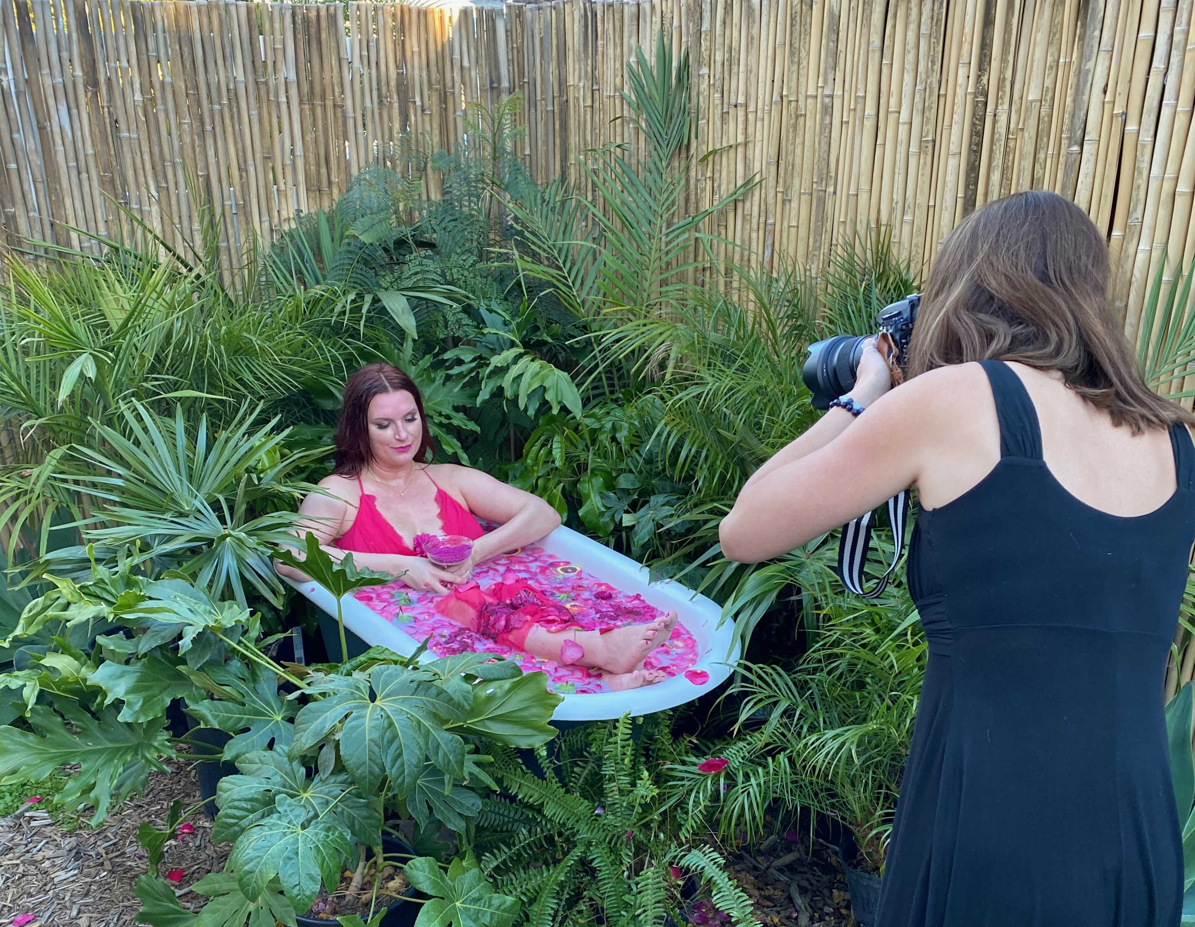 A woman in a bath surrounded by tropical plants poses for a photograph, while another woman in a black dress captures the moment with a camera.