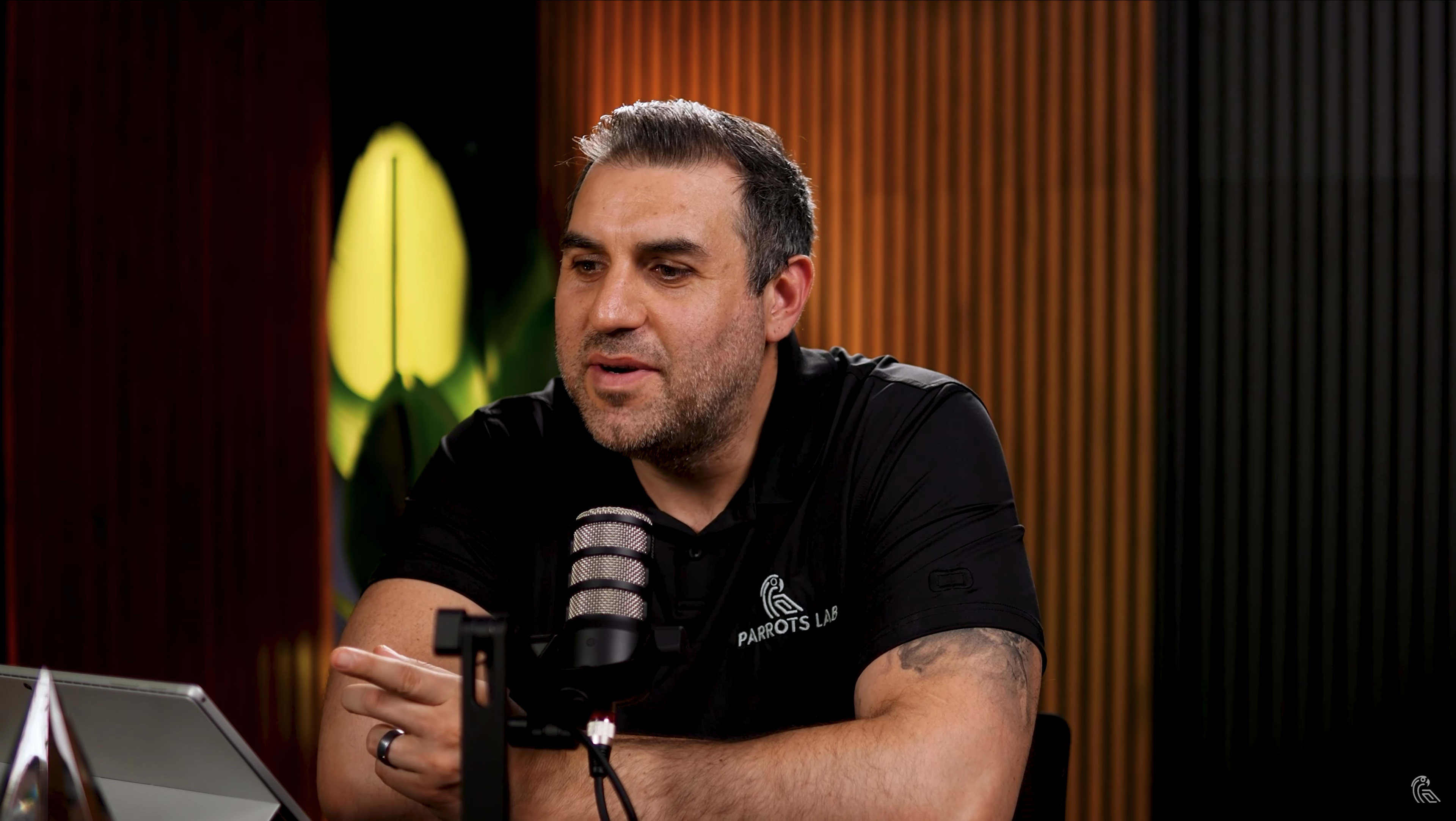 A man in a black shirt with a logo speaks into a microphone while seated at a table with a glass in front of him.