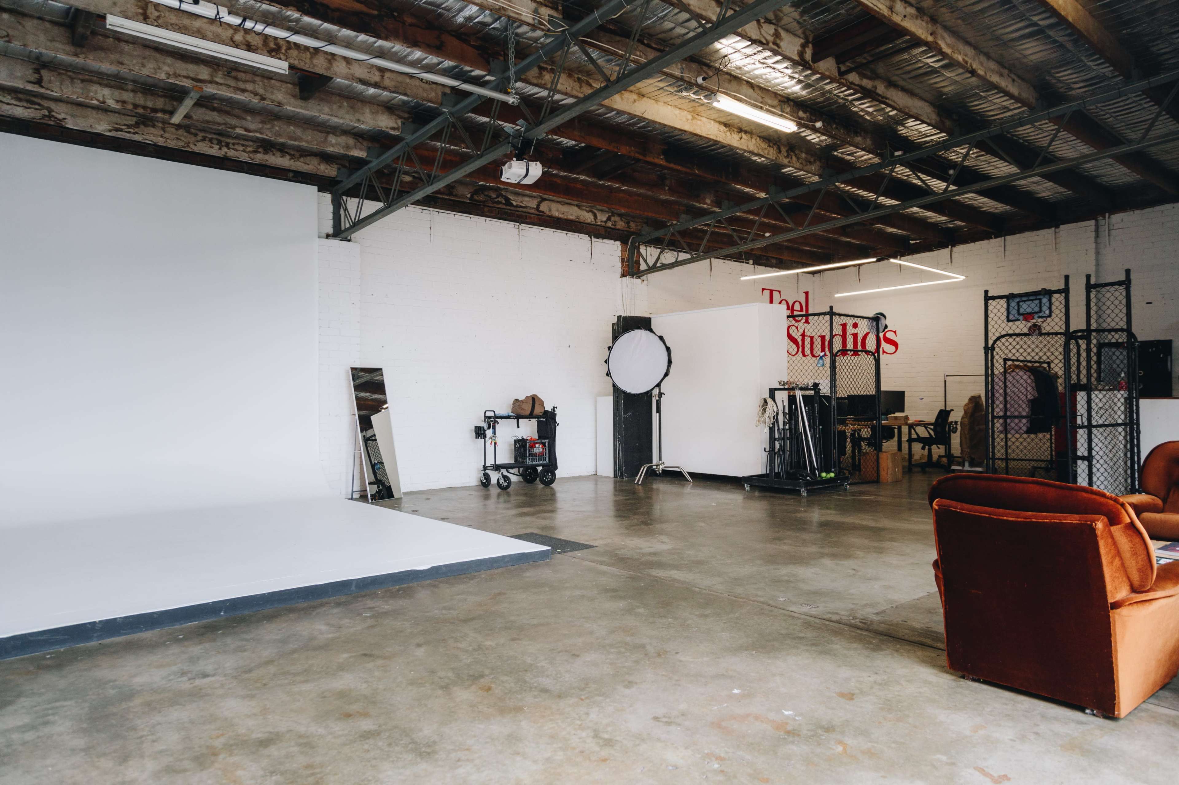 A spacious studio with a large white backdrop, studio equipment, and a brown couch against a concrete floor.