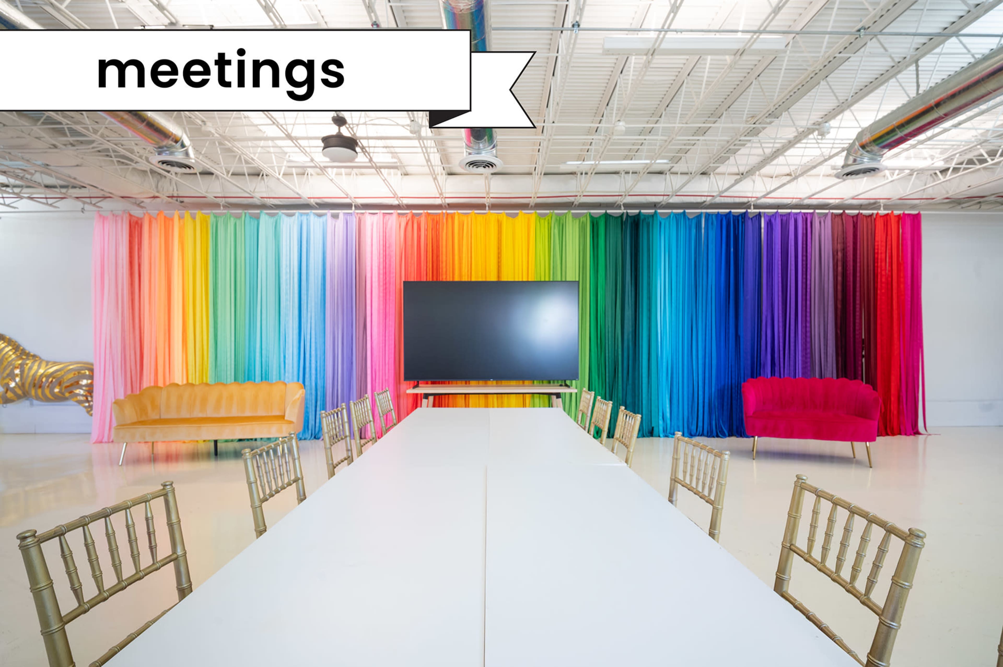 A long white table is set up in a brightly colored meeting room with a gradient backdrop and two distinct couches in vibrant colors.