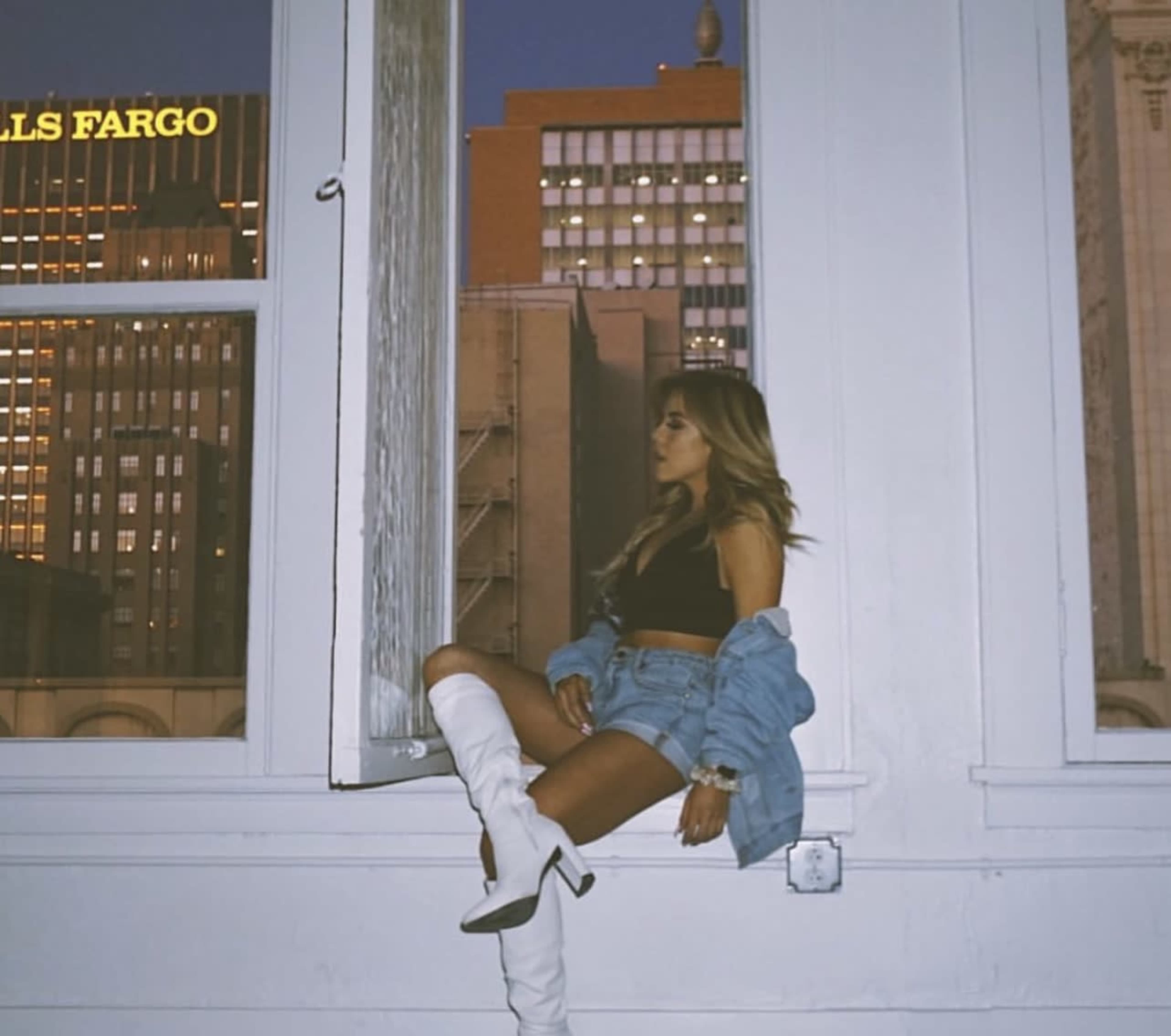 A woman sits on a ledge by an open window, wearing a black top, denim jacket, and white boots, with city buildings visible in the background.