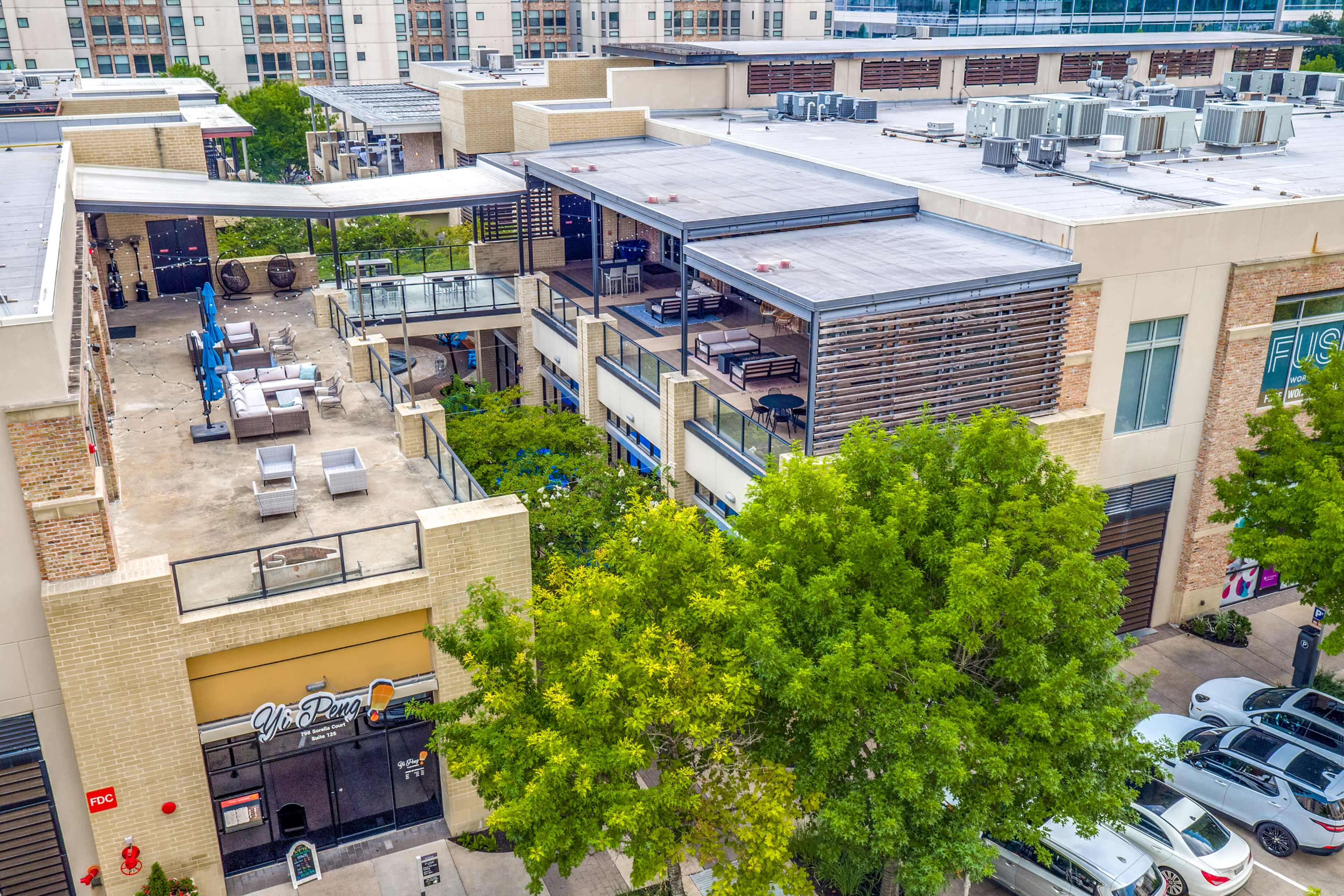 A multi-level commercial building with outdoor seating areas and air conditioning units on the roof, surrounded by trees and parked cars.