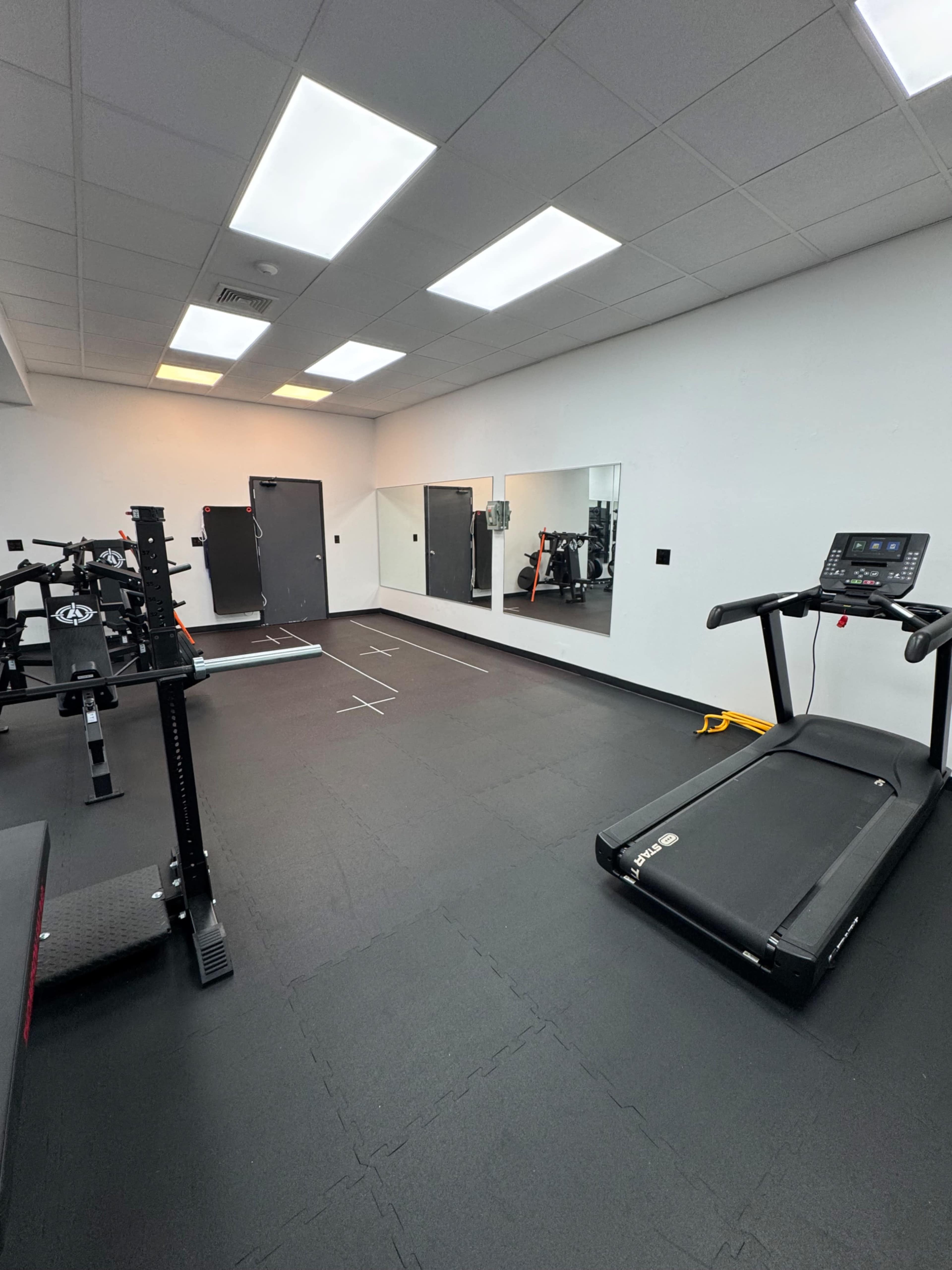The image shows a gym interior featuring a treadmill, a weightlifting rack, and mirrors on the walls.