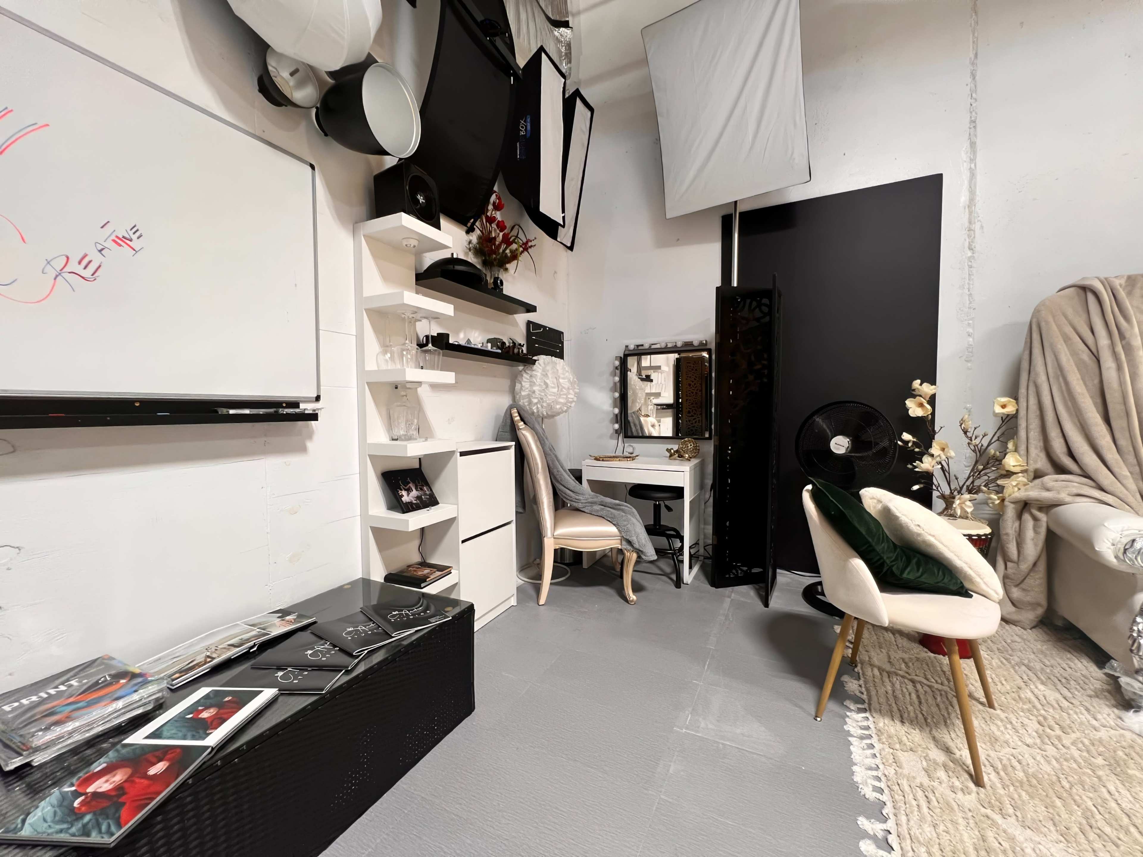 A neatly organized studio space featuring a whiteboard, shelves with decorative items, a dressing table, seating, and a backdrop against a wall.