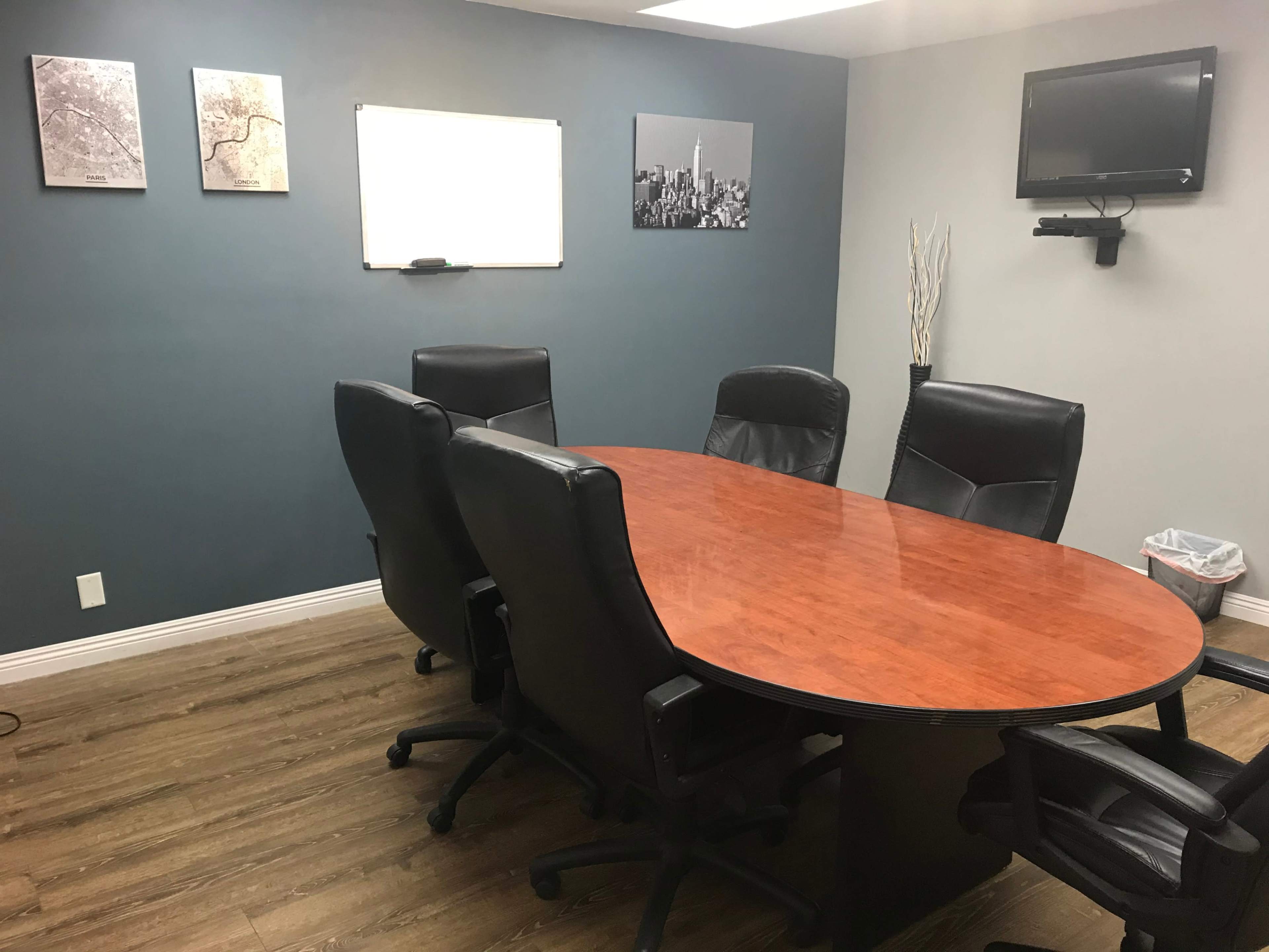 A conference room features an oval table surrounded by six black chairs, with a whiteboard and two framed images on the wall.