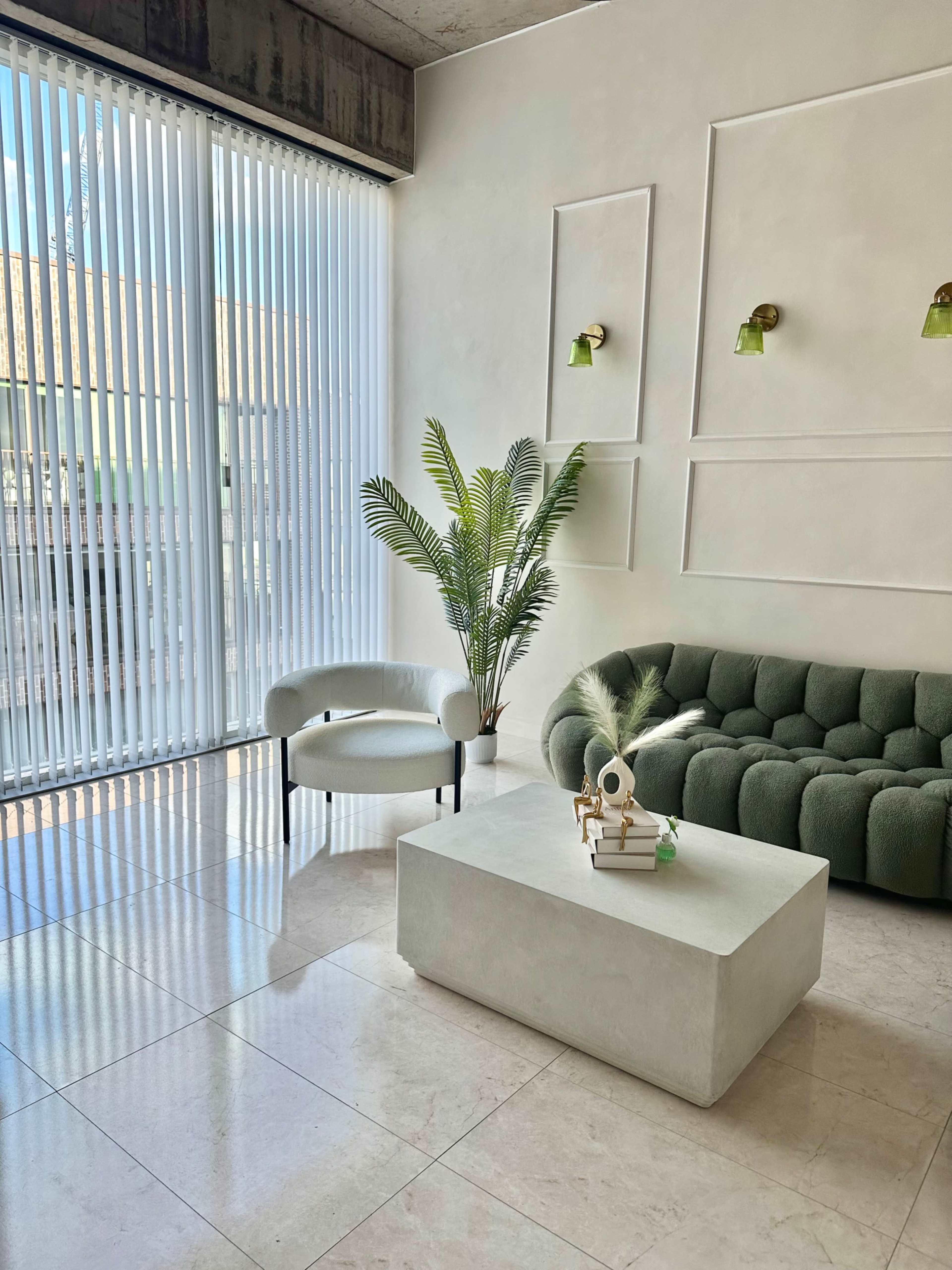 The image depicts a modern living room with a green sofa, a white armchair, and a rectangular coffee table, illuminated by natural light from a large window with vertical blinds.