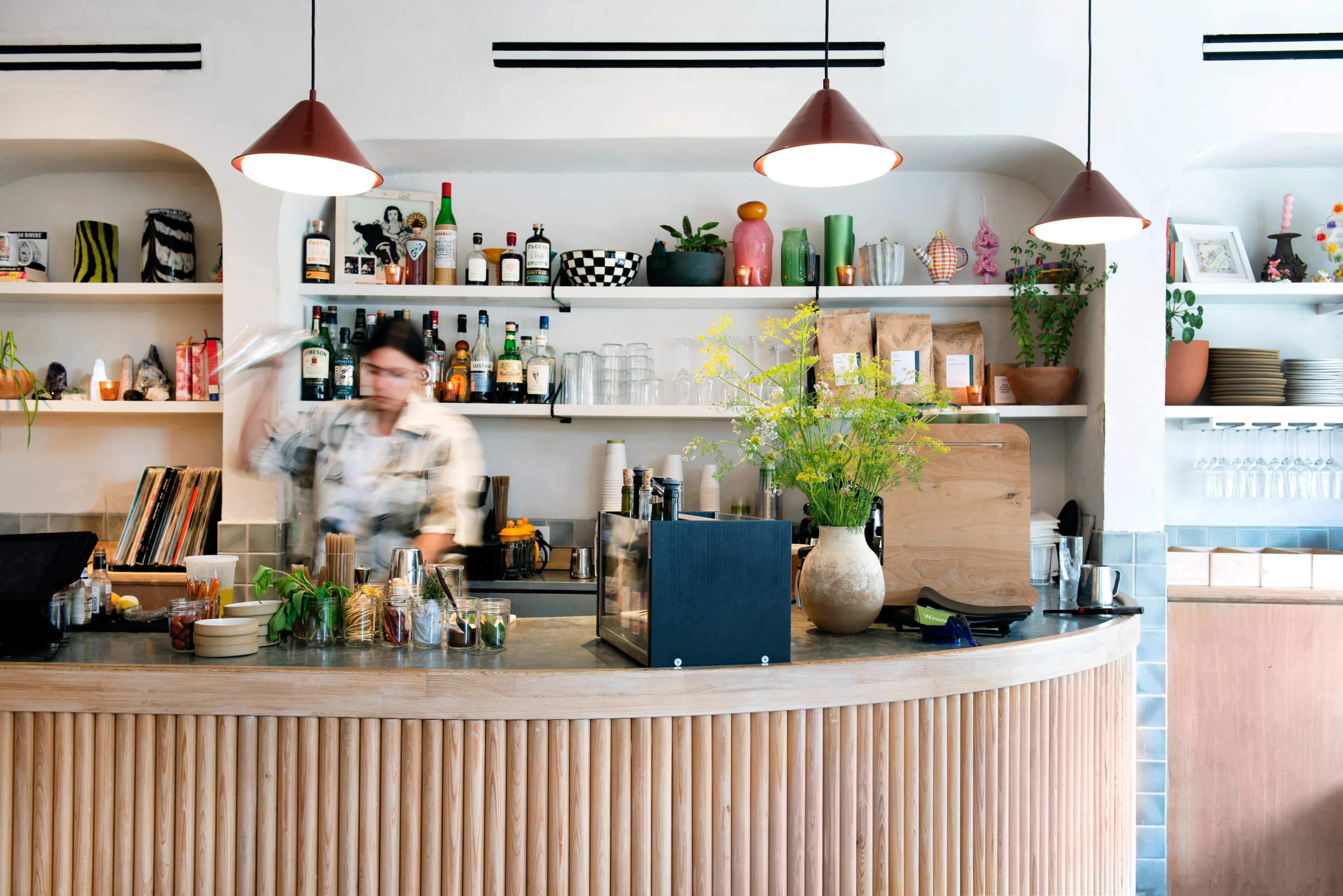 A bartender prepares drinks at a curved wooden bar adorned with various bottles and decorative plants in a brightly lit café.