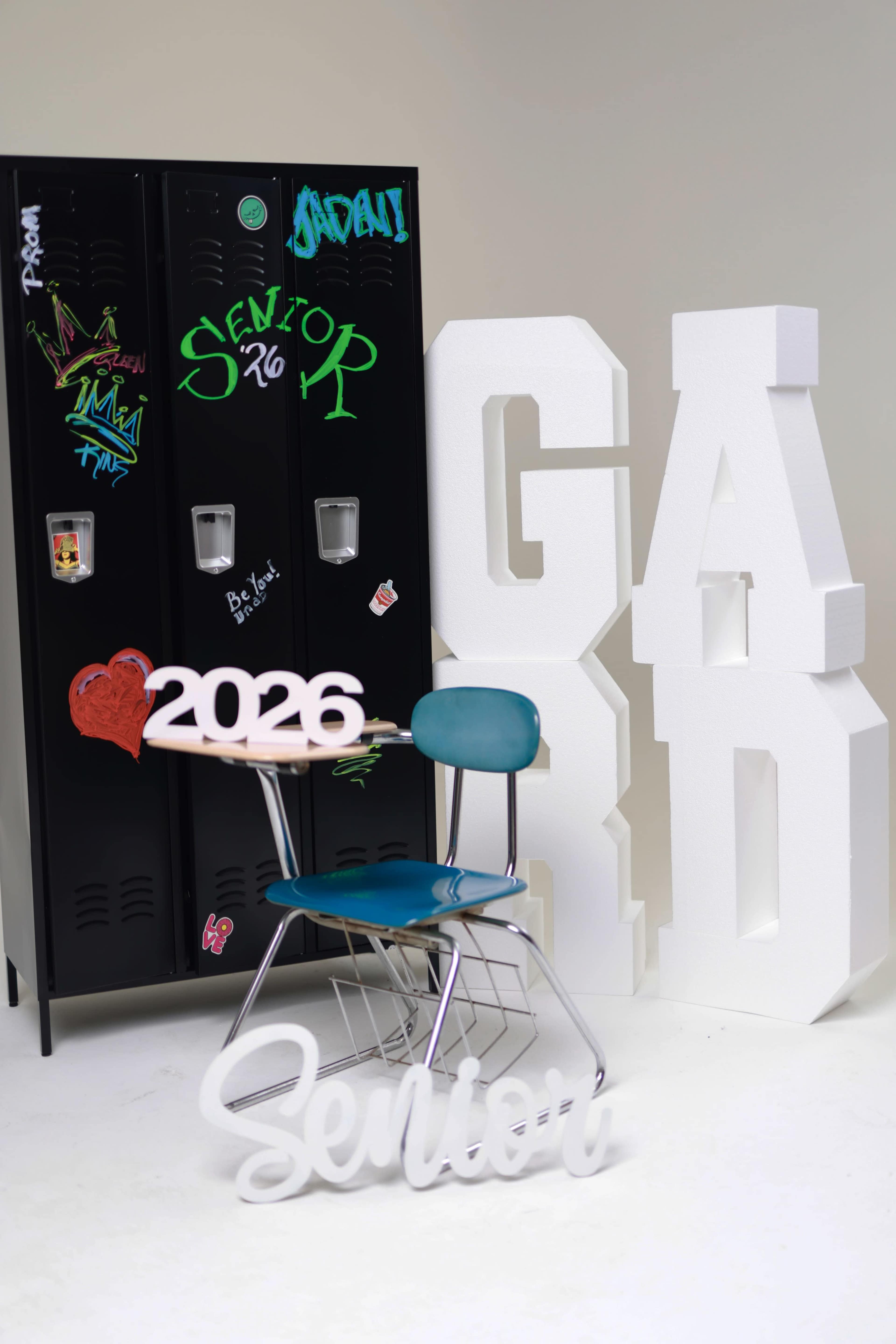 The image features a blue school chair positioned in front of black lockers, alongside large white foam letters spelling "GRAD" and "2026," with the word "Senior" displayed prominently on the floor.