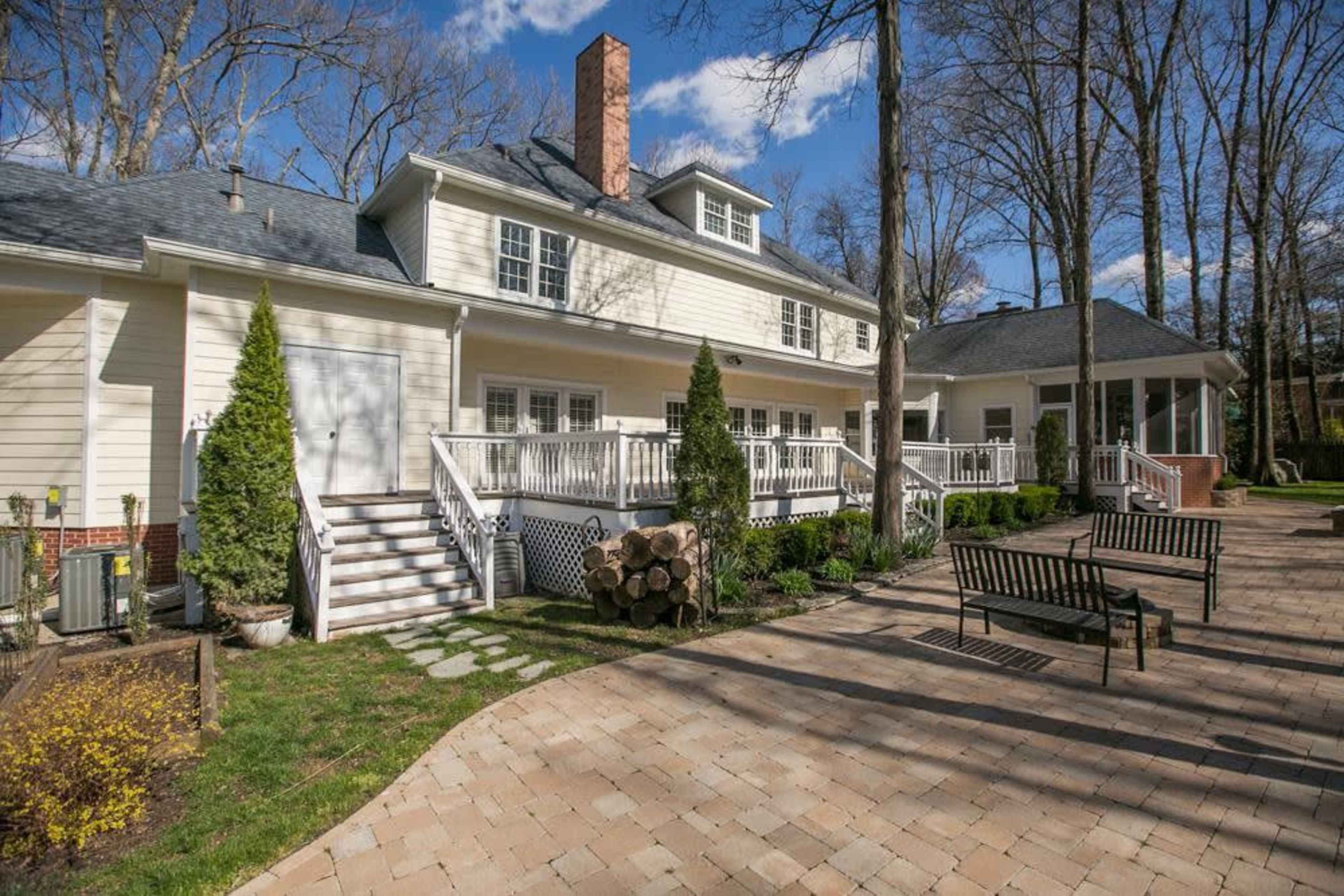 A large, light-colored house with a sloped roof, a brick chimney, a spacious deck, and a paved patio area surrounded by trees.