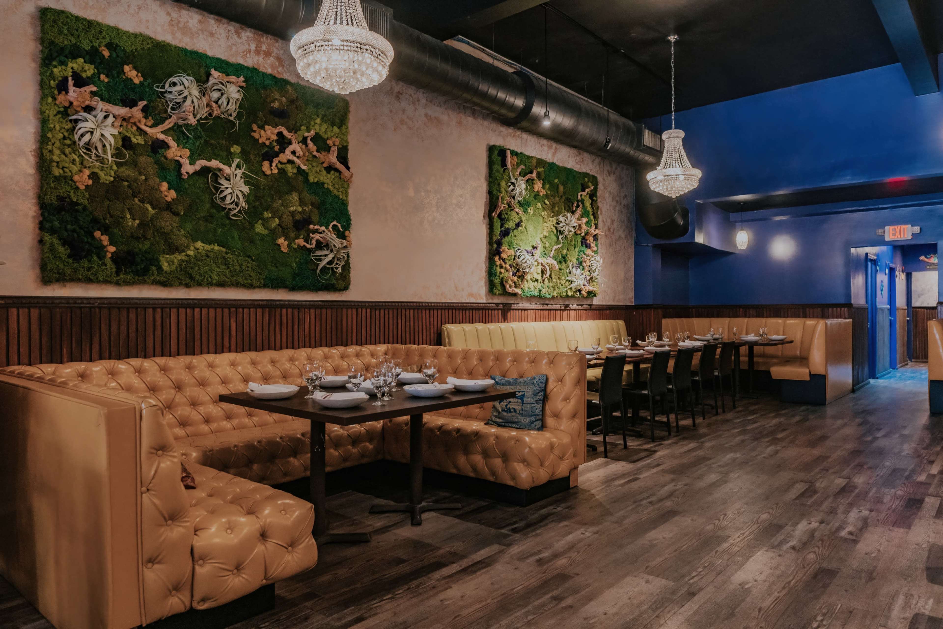 The image shows a modern restaurant interior featuring a yellow tufted booth, wooden paneling, and two large decorative wall pieces with greenery.