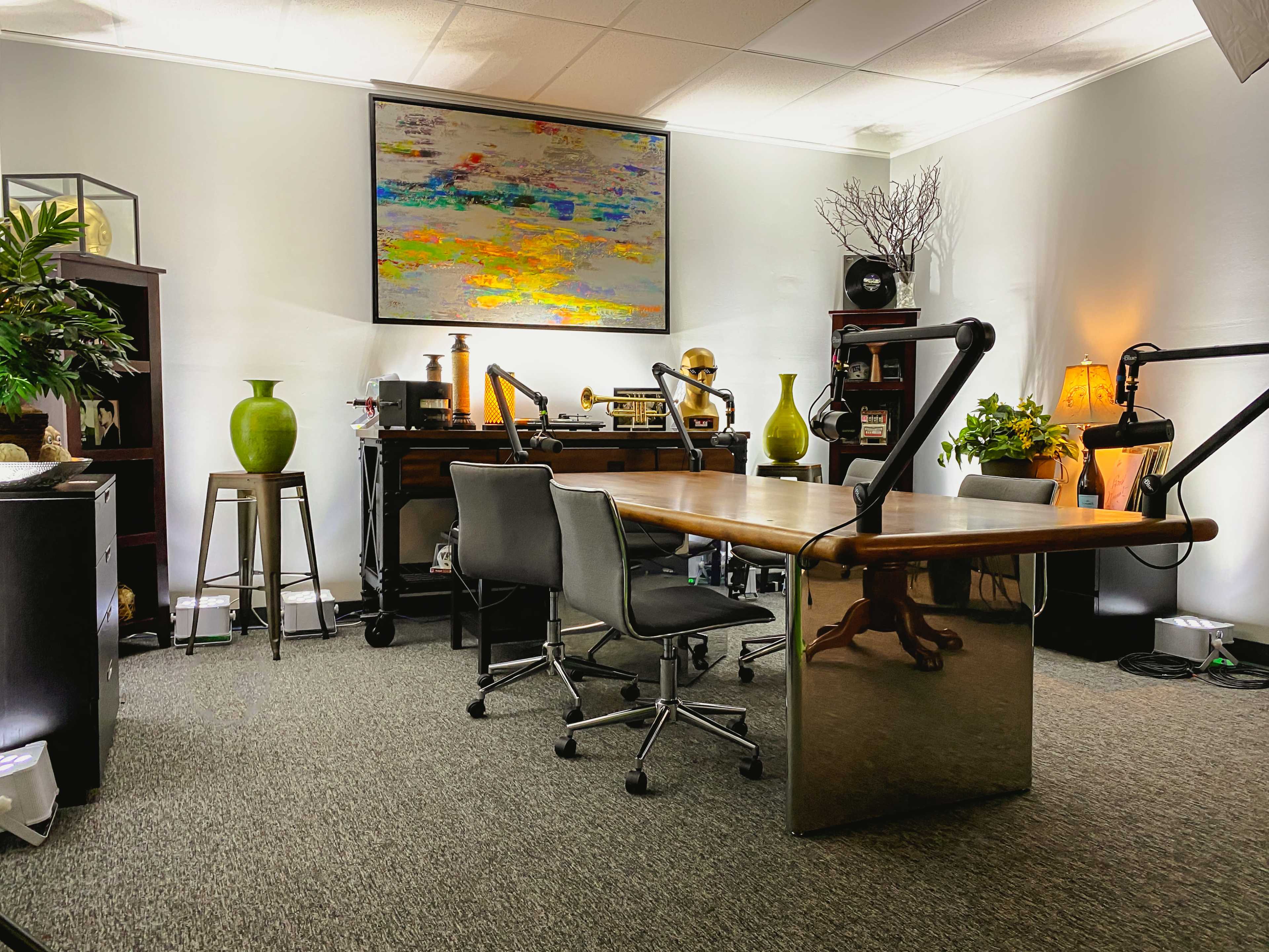 A modern office space features a large wooden table with two chairs, surrounded by plants, decor, and a colorful painting on the wall.