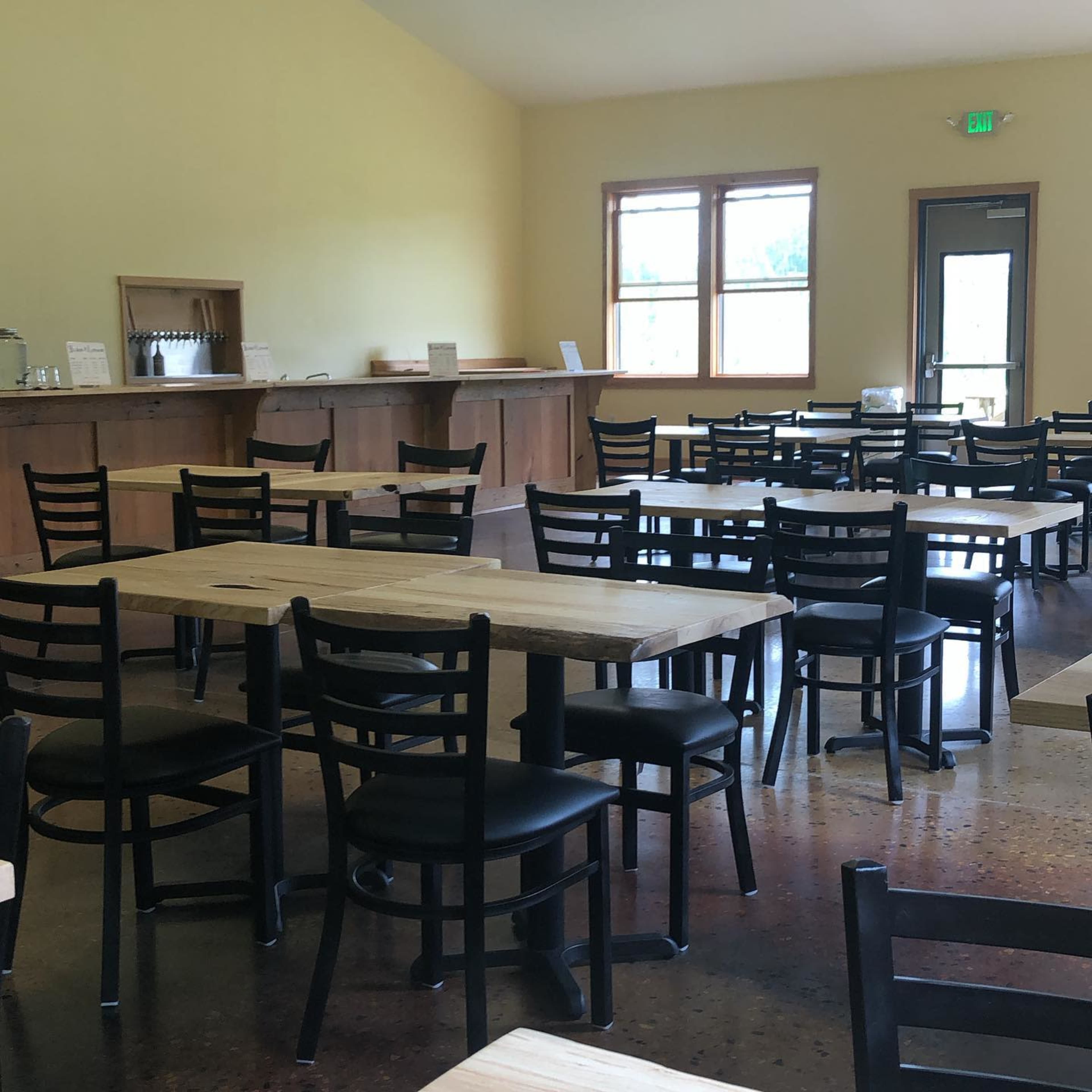 The image shows a spacious dining area with several wooden tables and black chairs, illuminated by natural light from windows.