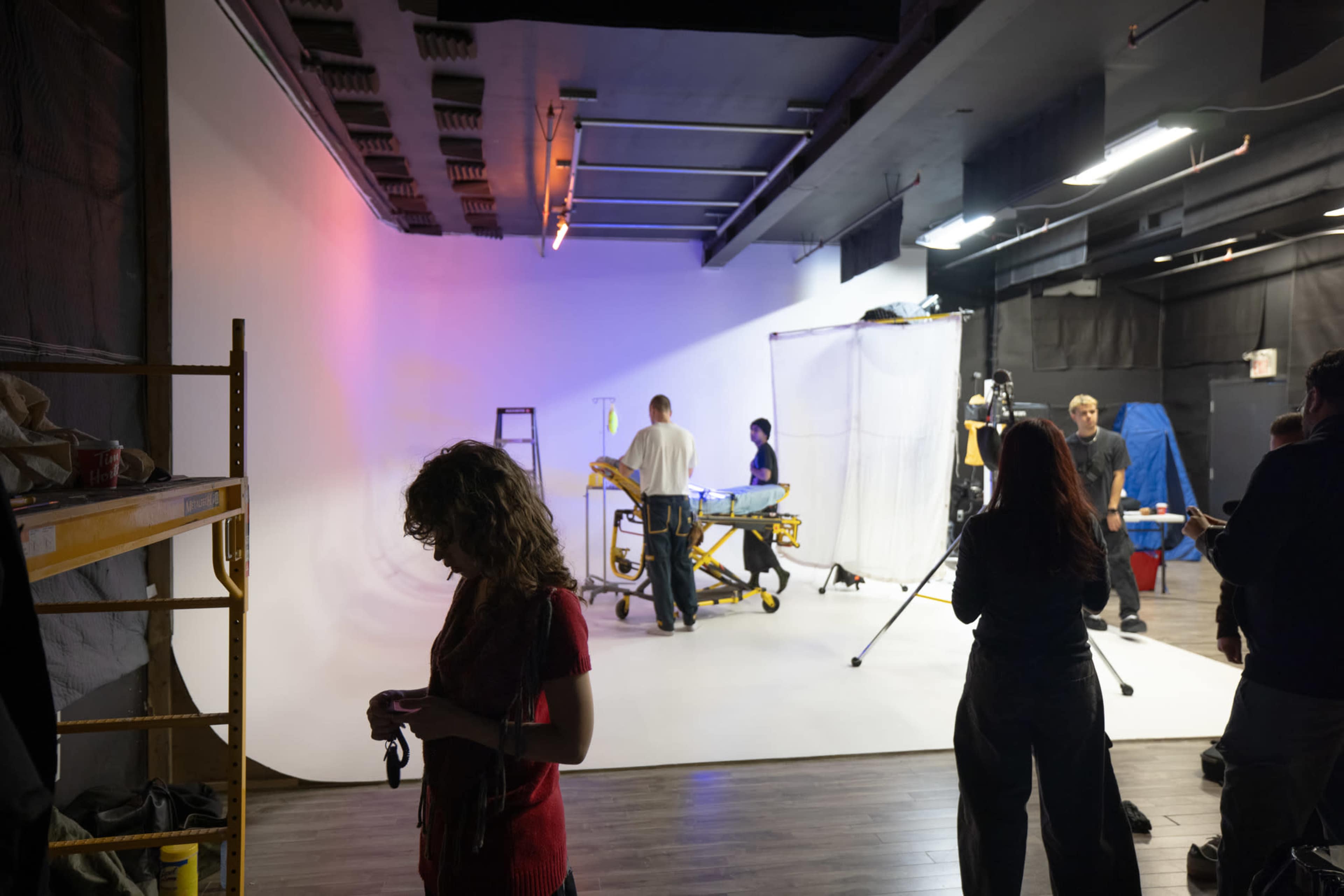 A film studio scene features several people working on a set with a lit backdrop, equipment, and props arranged in the space.