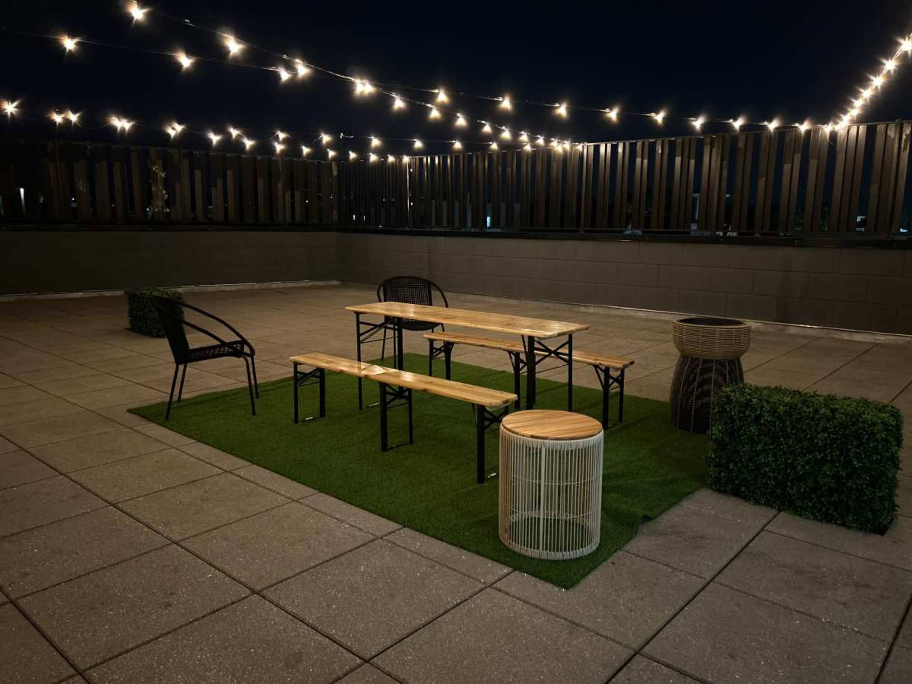 A rooftop space features a wooden table and benches set on artificial grass, surrounded by string lights and various seating options.