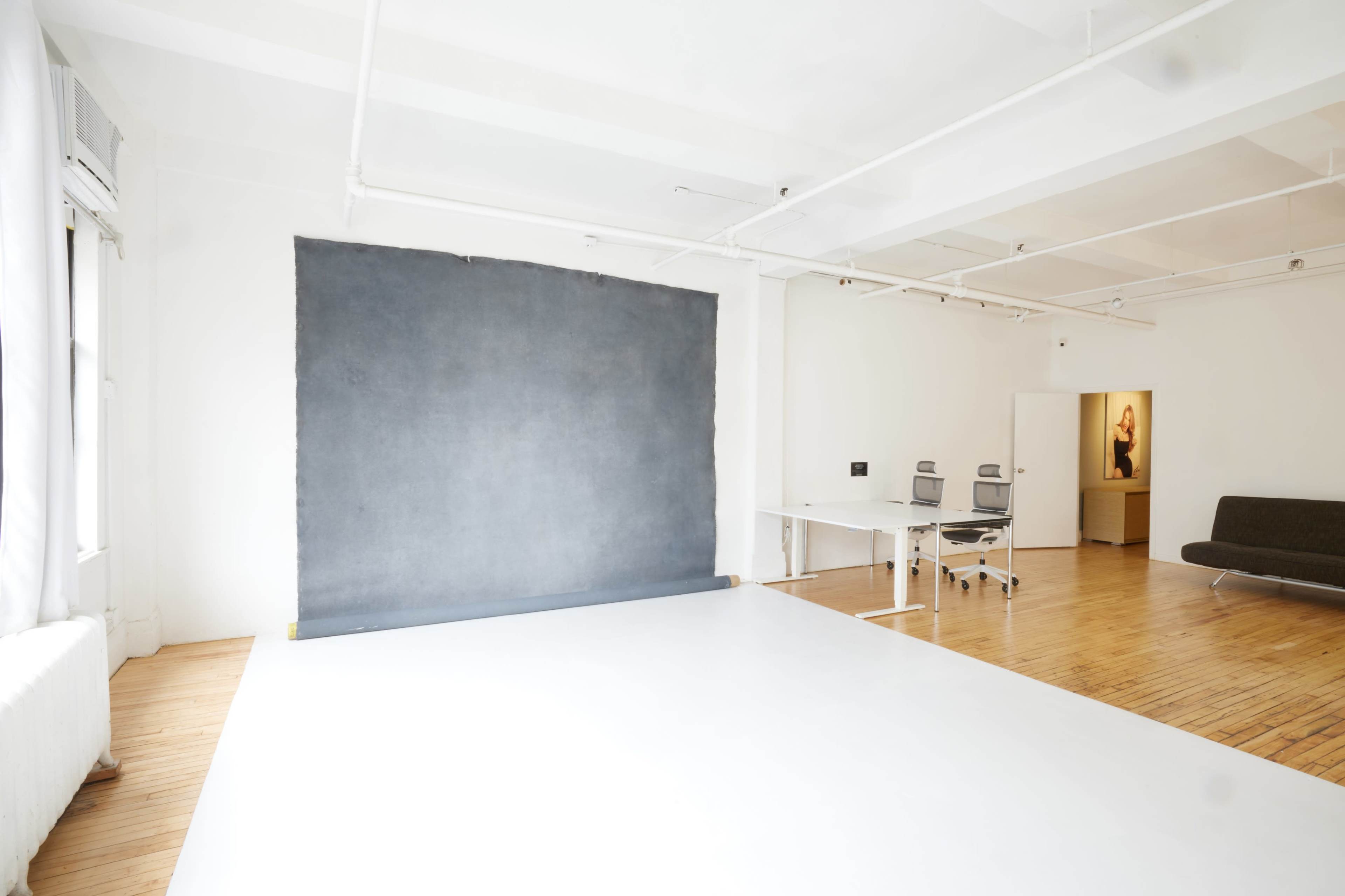 A spacious, minimalist studio with a large gray backdrop against one wall, a desk with office chairs in the center, and a sofa along another wall.