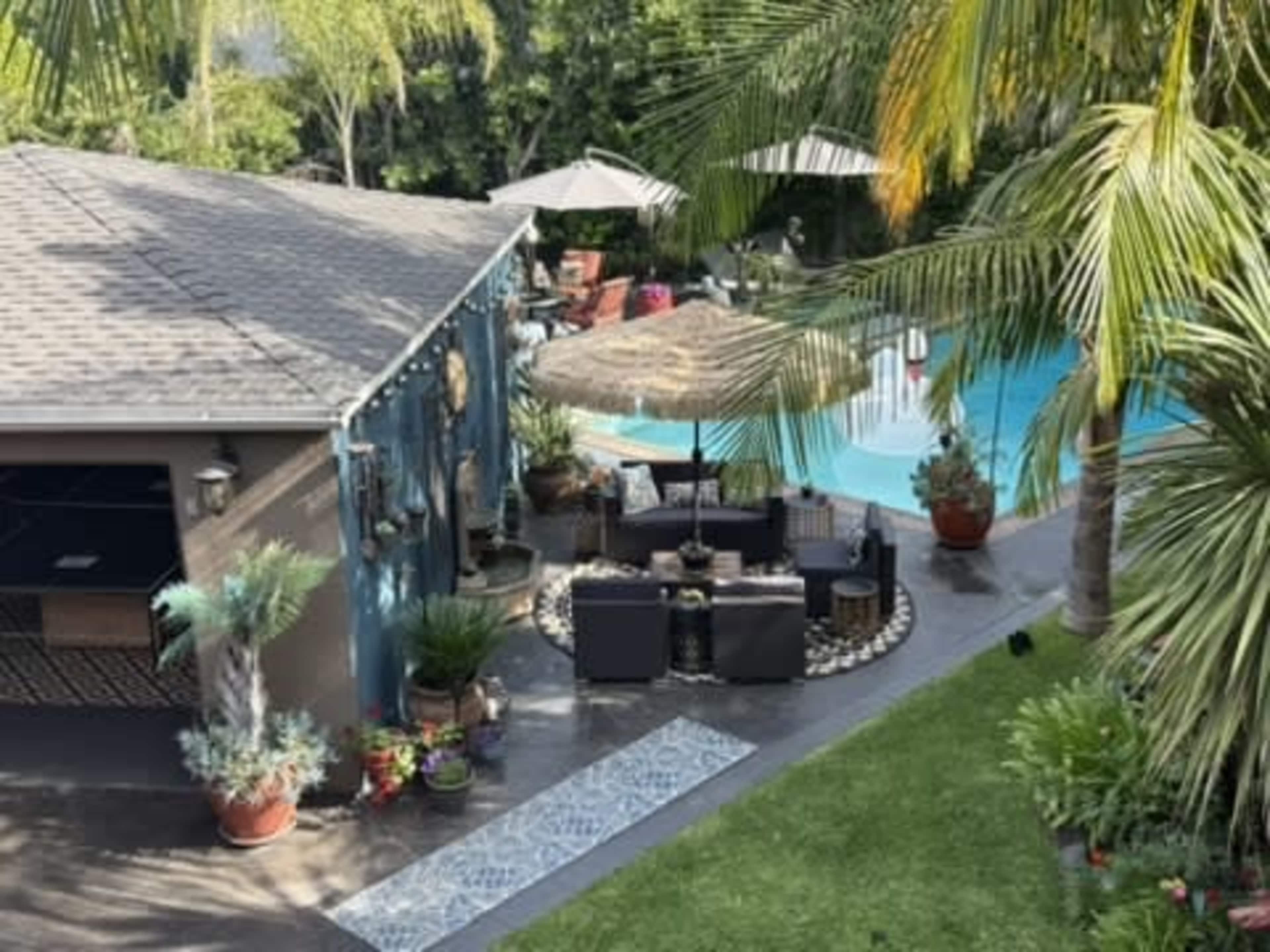 The image shows a backyard with a swimming pool, a shaded seating area surrounded by potted plants, and several umbrellas.