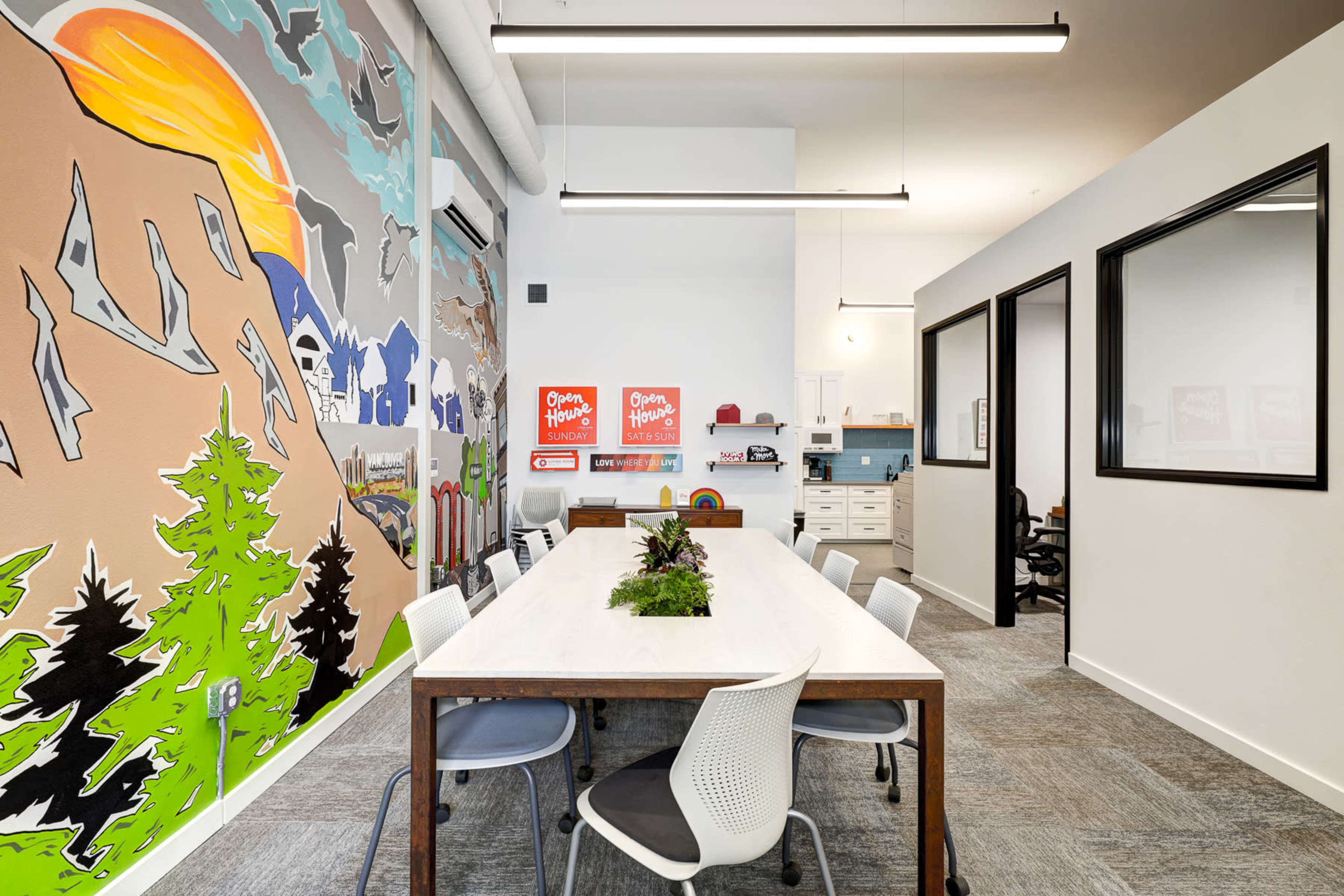 A modern office space features a large table surrounded by chairs, with a colorful mural on one wall and organized workspaces in the background.