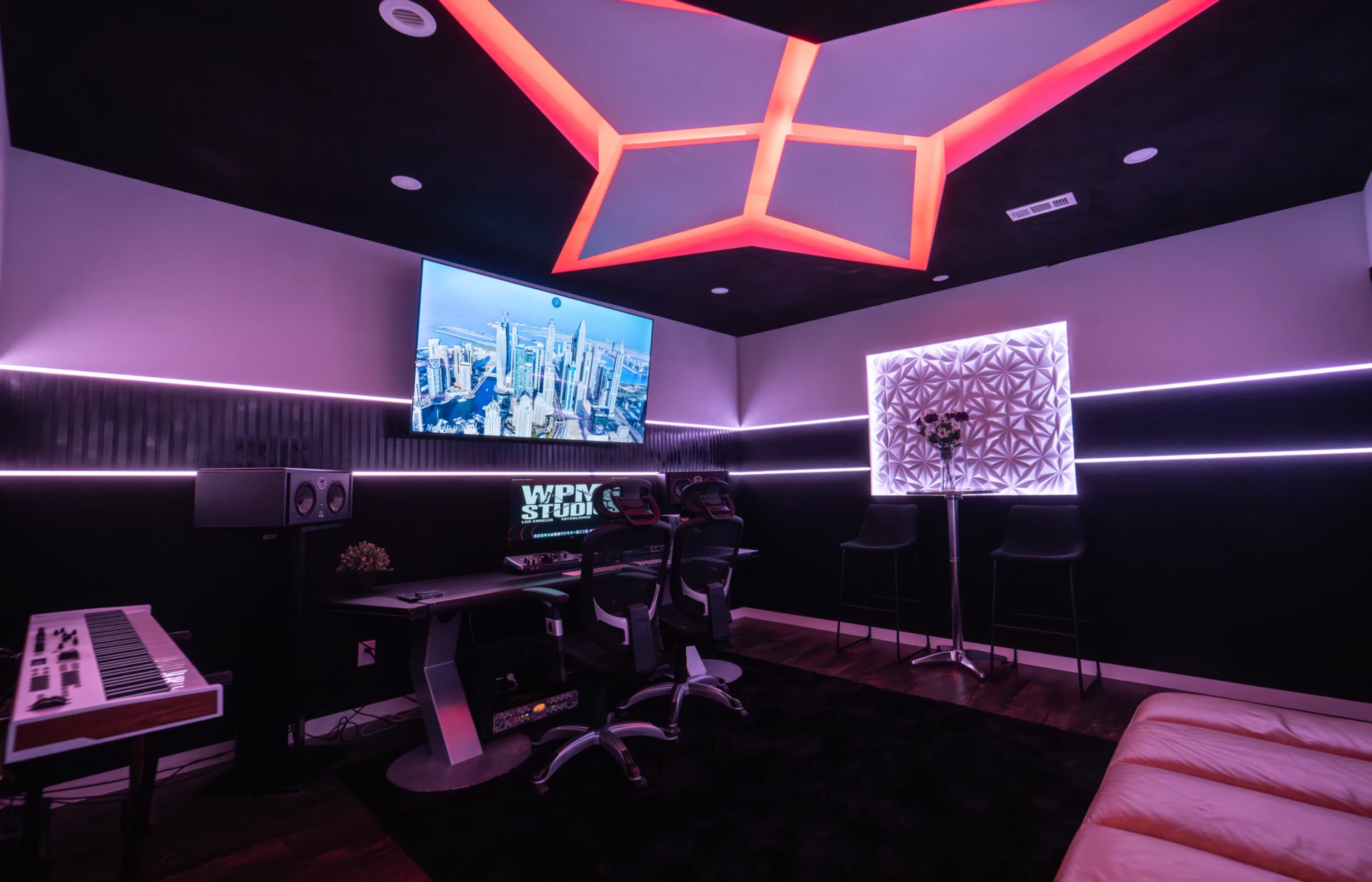 The image shows a modern studio with a large TV mounted on the wall, vibrant lighting, a desk with computers, and seating areas.