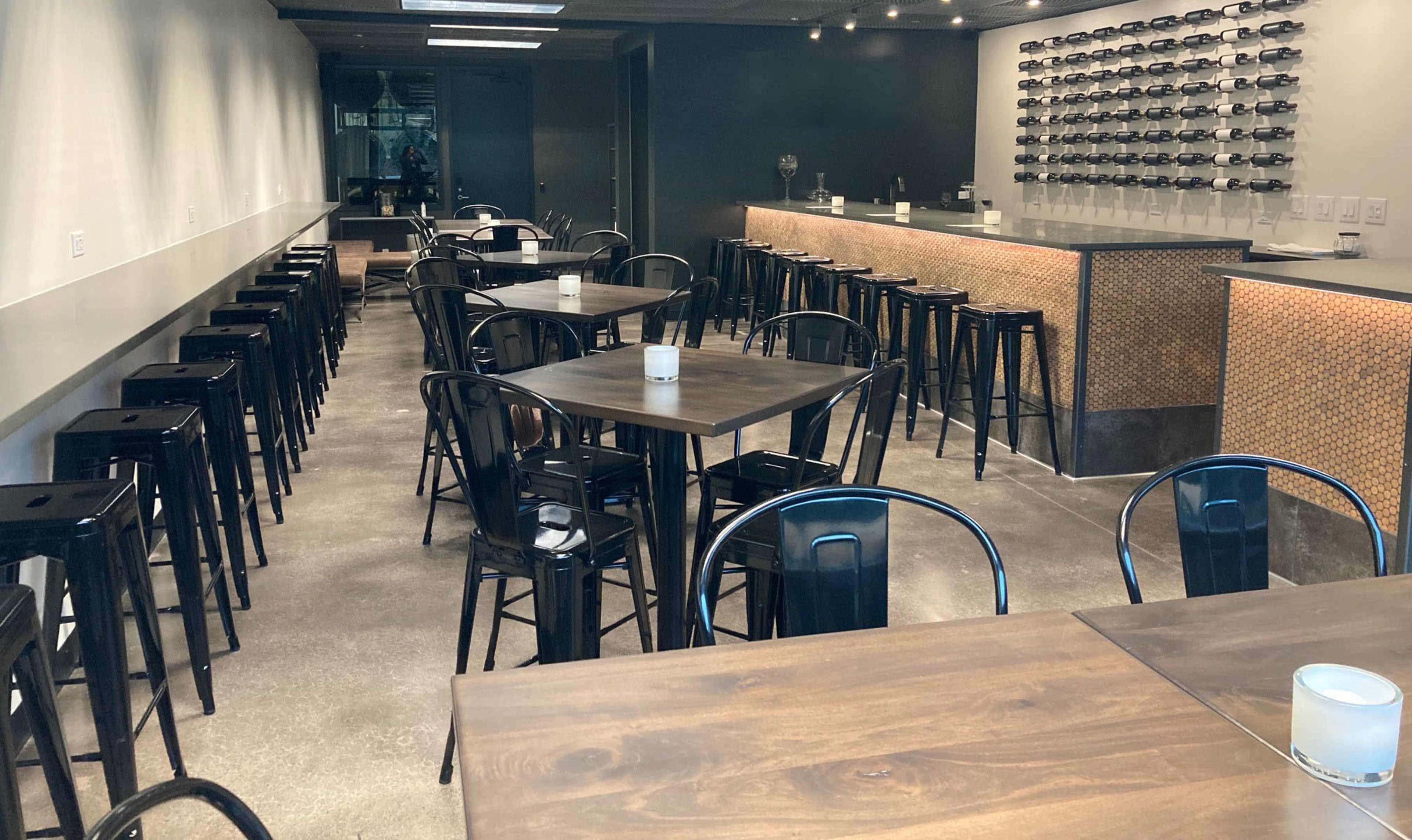 A modern dining area with several wooden tables, black metal stools, and a bar featuring a wall of wine bottles.