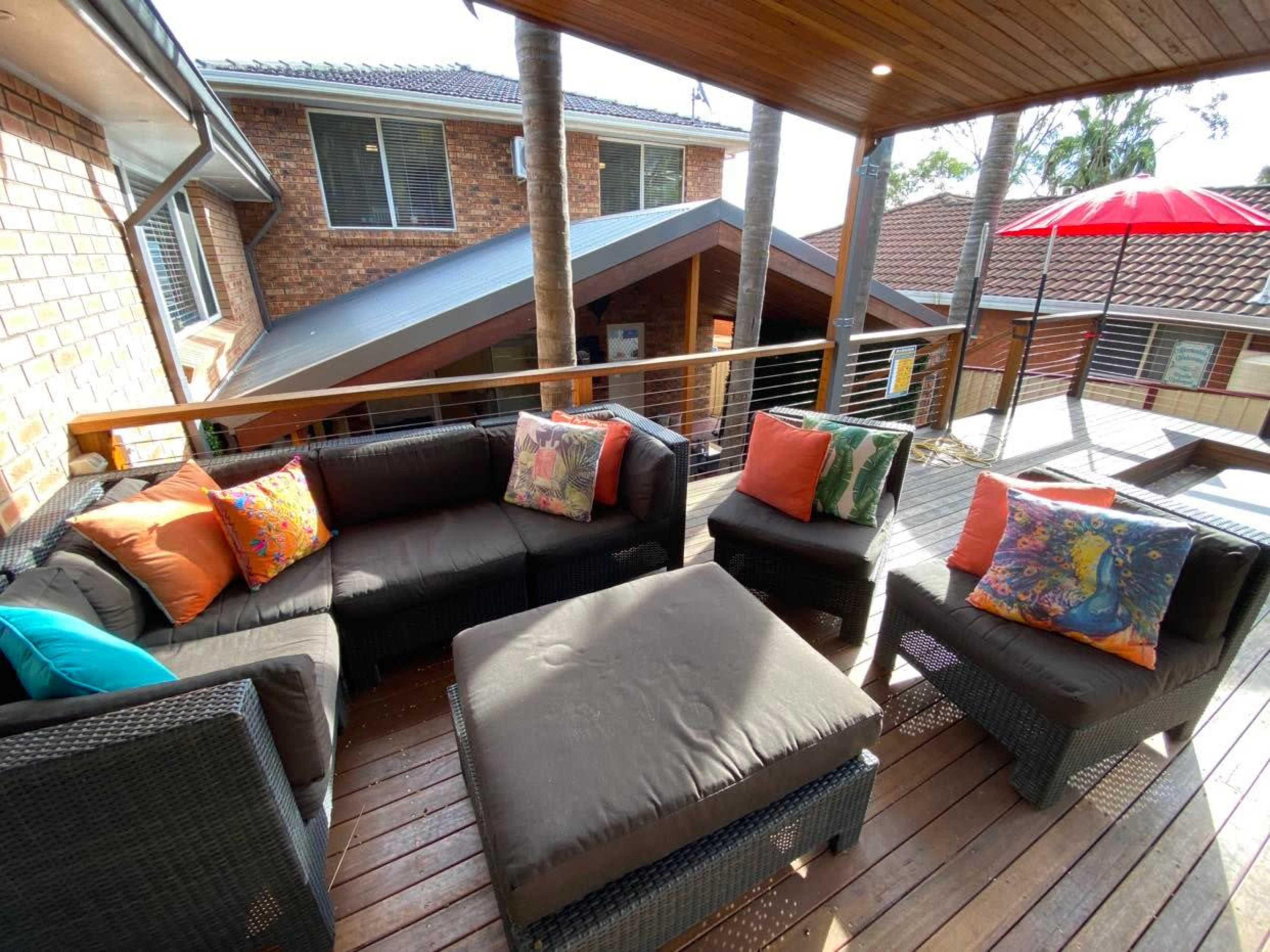 A covered outdoor seating area with a wicker couch, colorful pillows, and a square coffee table on a wooden deck.