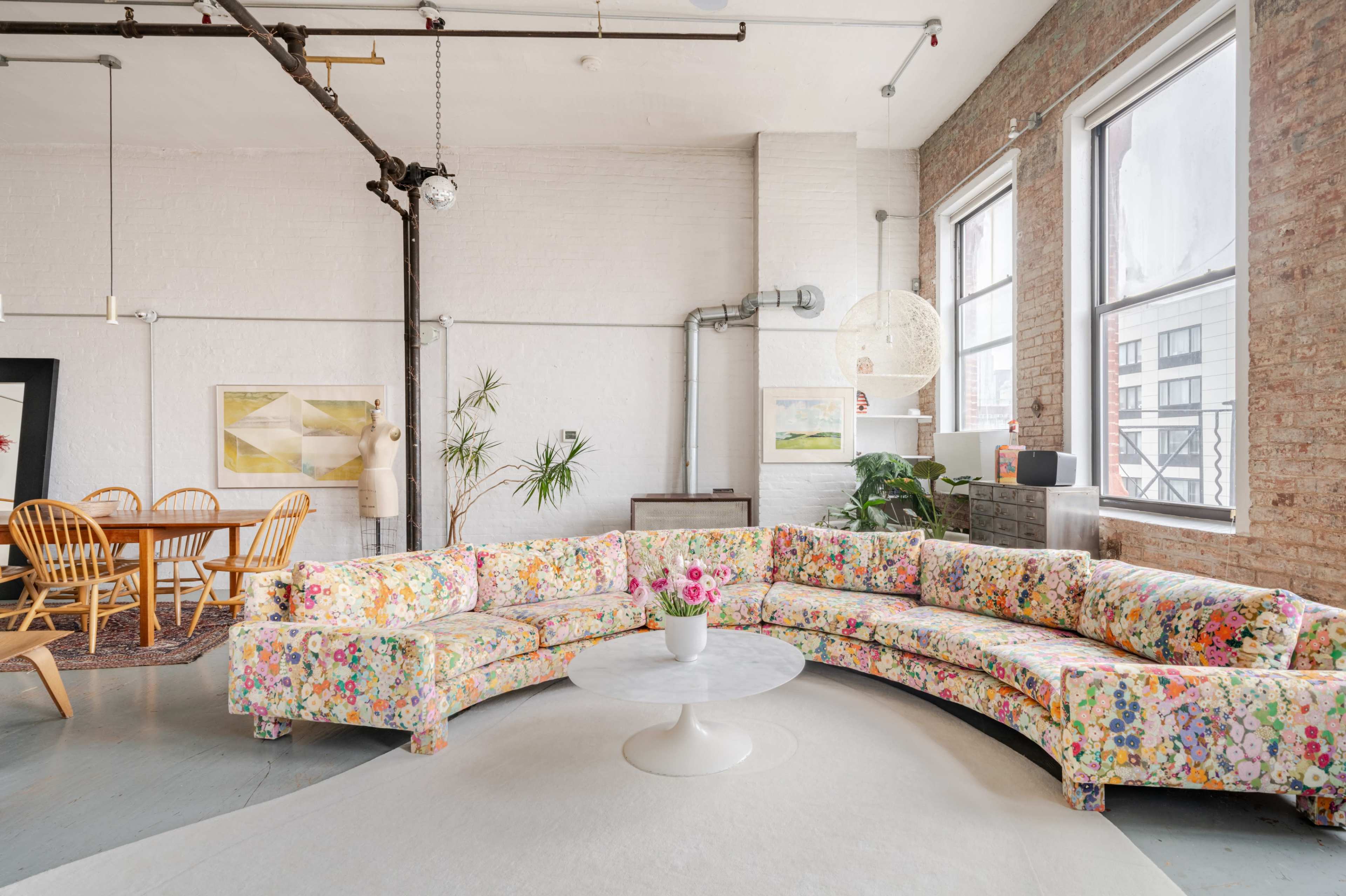 The image depicts a bright and airy living space featuring a large, floral-patterned sectional sofa, a round coffee table, and a dining area with wooden chairs and a rug.