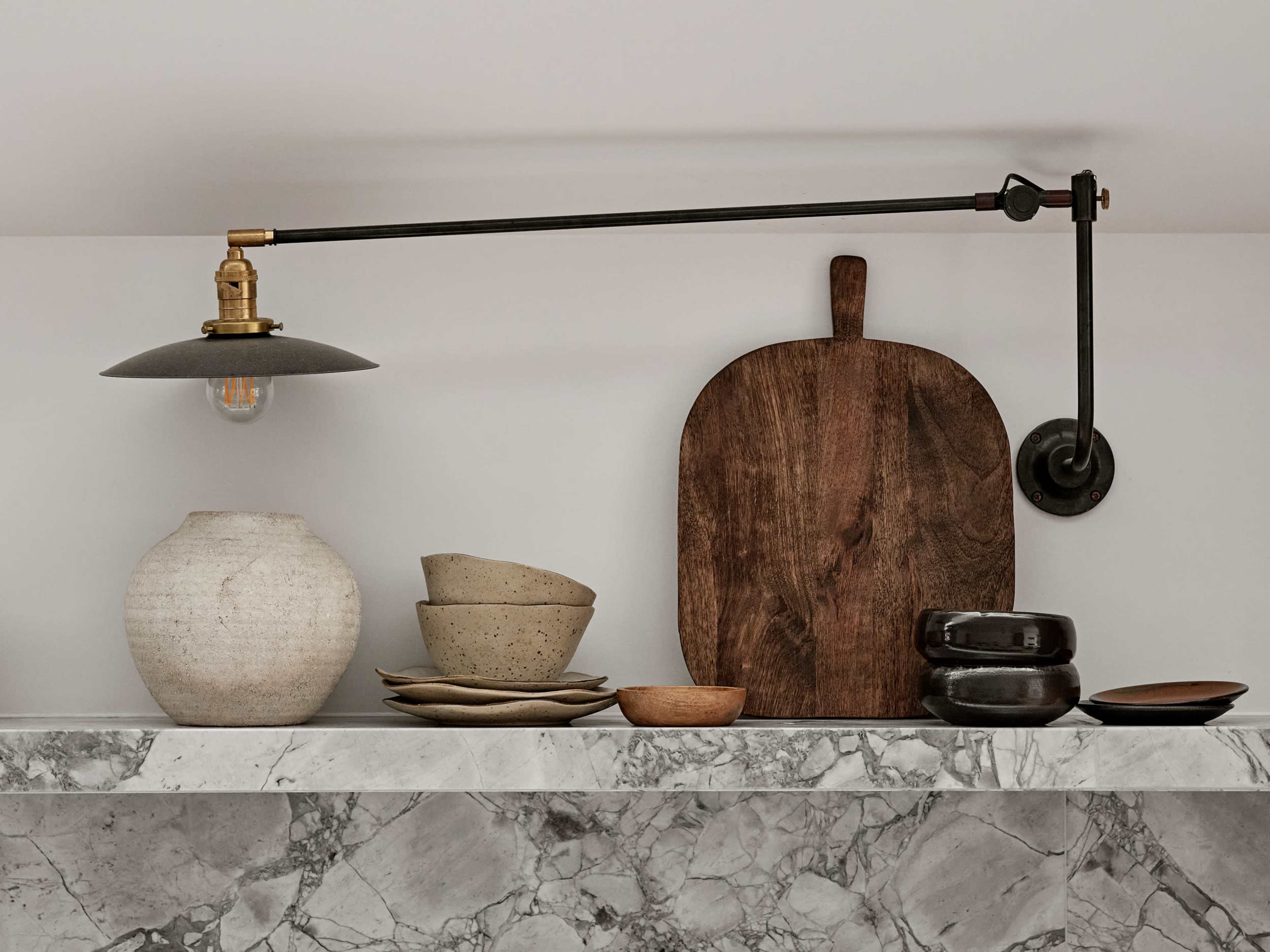 A wall-mounted lamp is positioned above a shelf displaying various ceramic bowls and wooden serving platters.