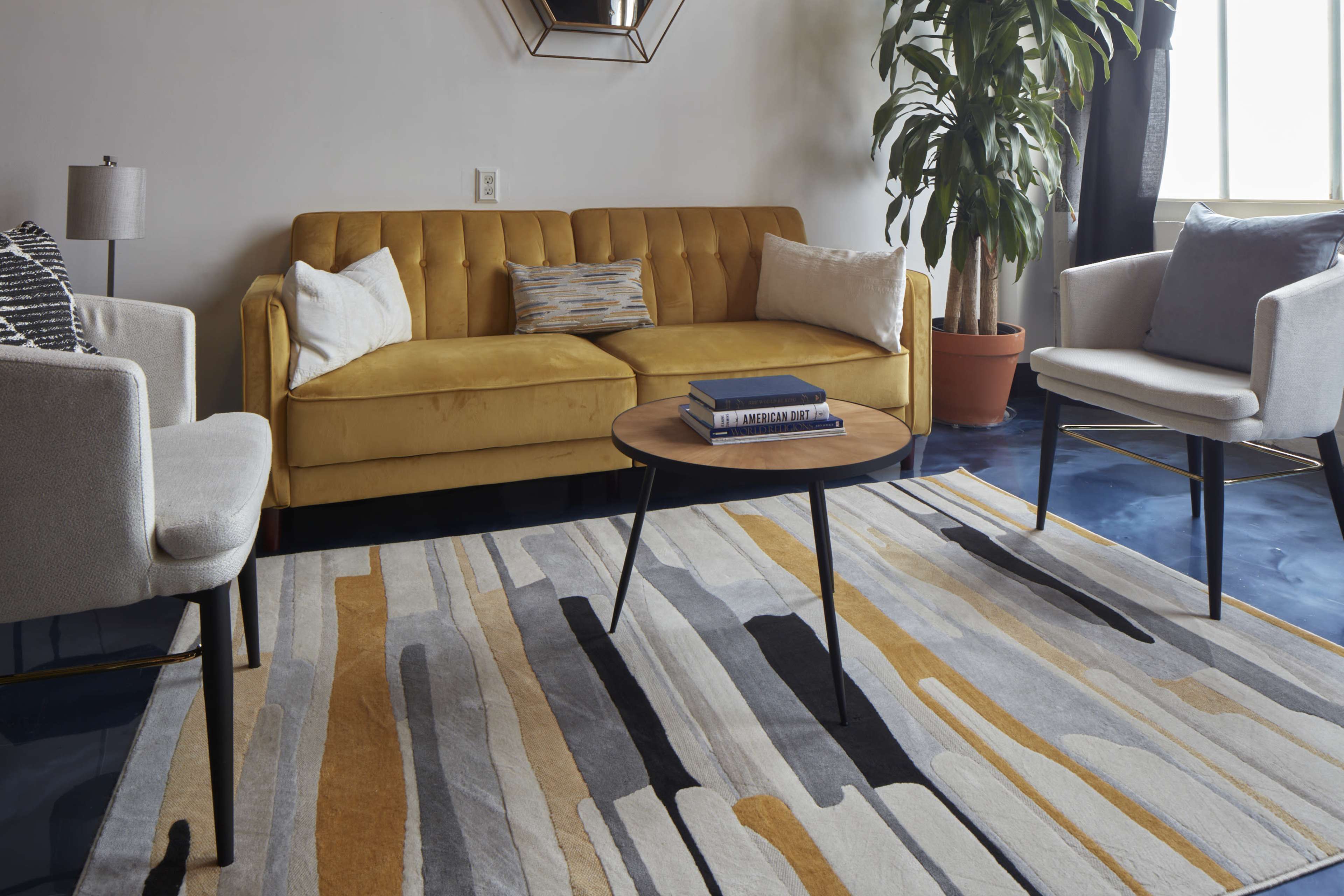 A yellow tufted sofa is paired with two light-colored chairs and a round coffee table on a striped area rug in a bright living room.