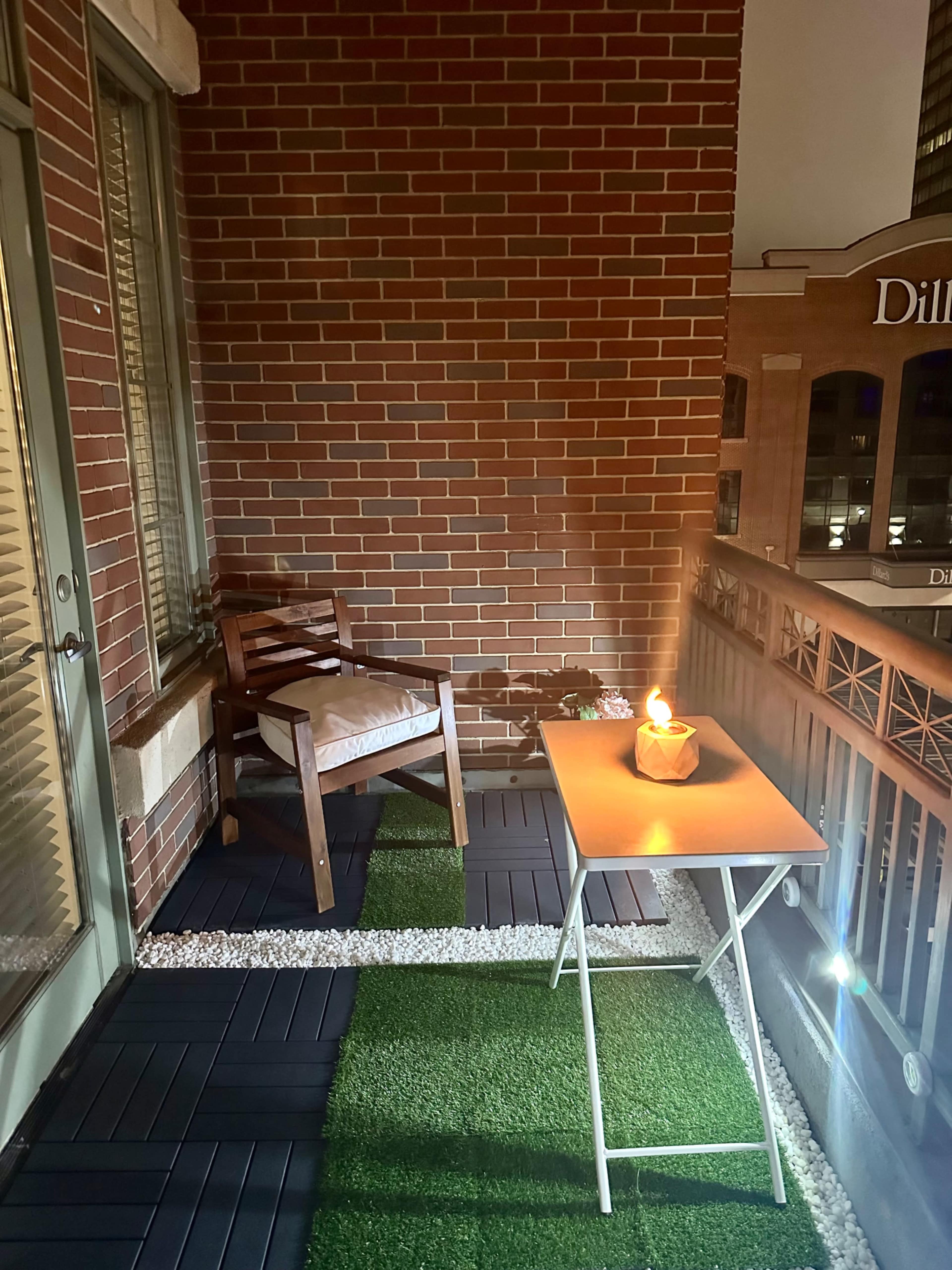 A small balcony with a wooden chair and a folding table, both illuminated by a candle, set against a backdrop of brick walls and a cityscape.