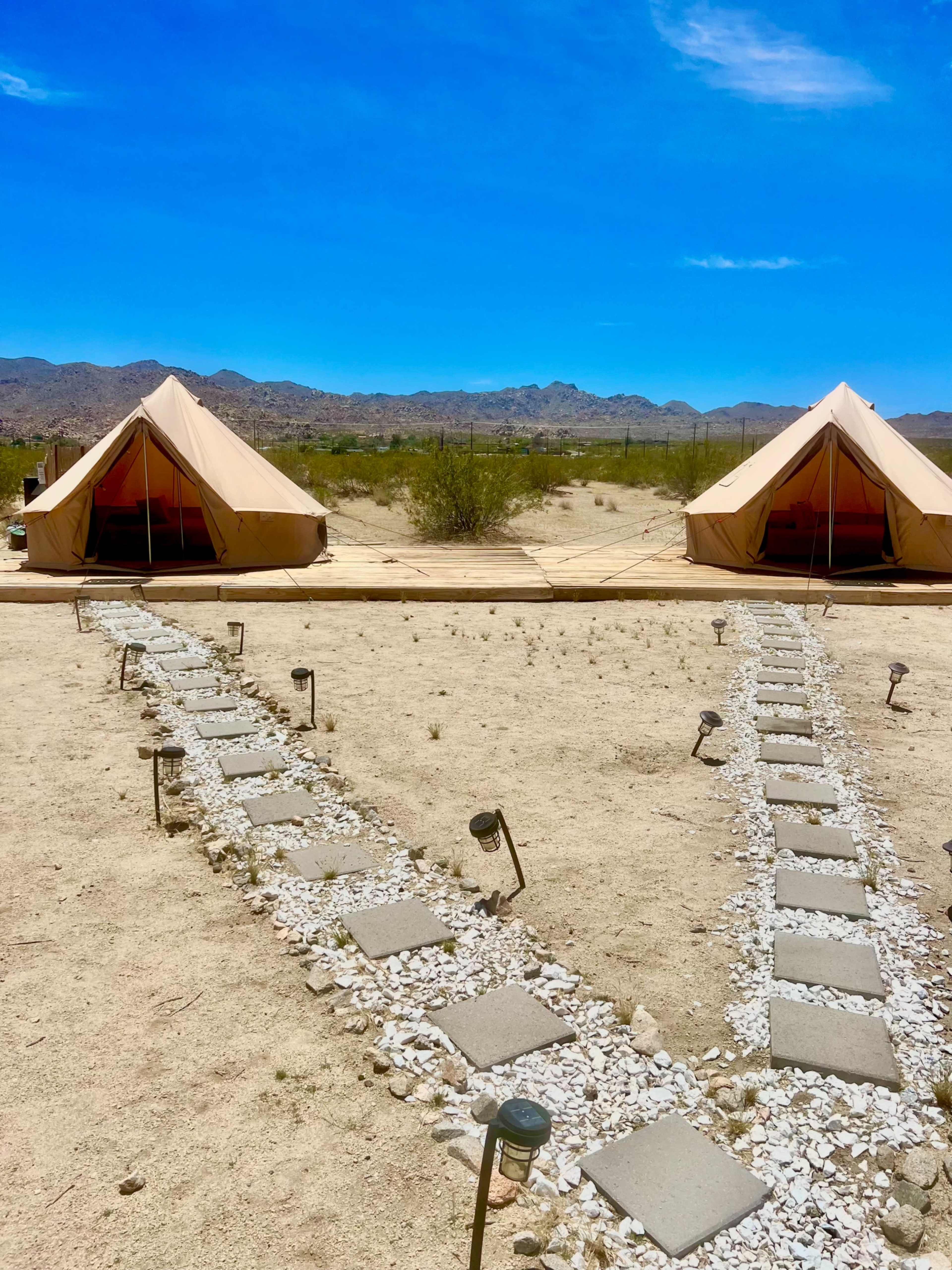 Two tents are positioned on a dirt pathway lined with stepping stones and small lights, set against a backdrop of low mountains under a clear blue sky.