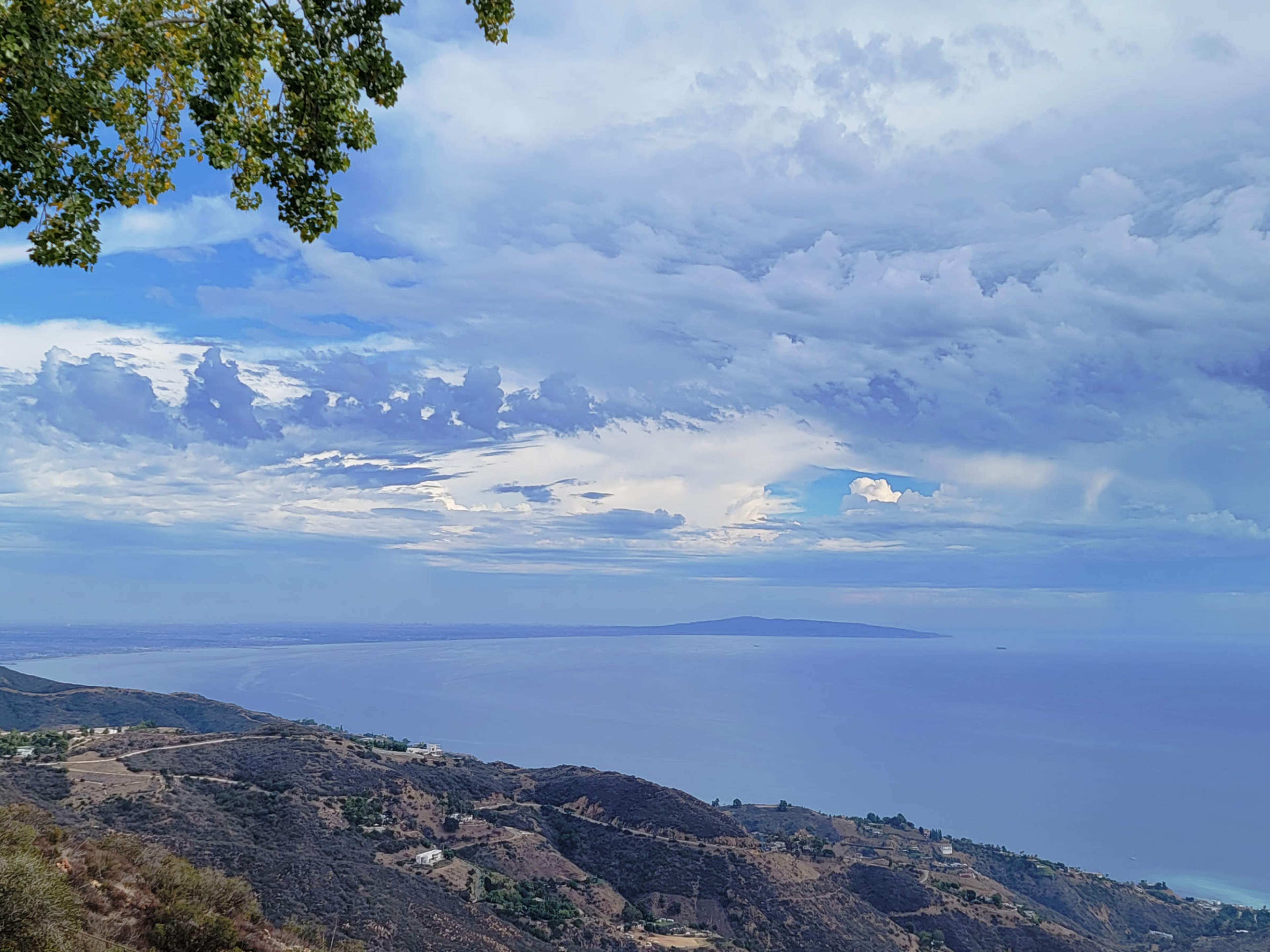 The image shows a panoramic view of a coastal landscape with rolling hills, a calm ocean, and a cloudy sky.