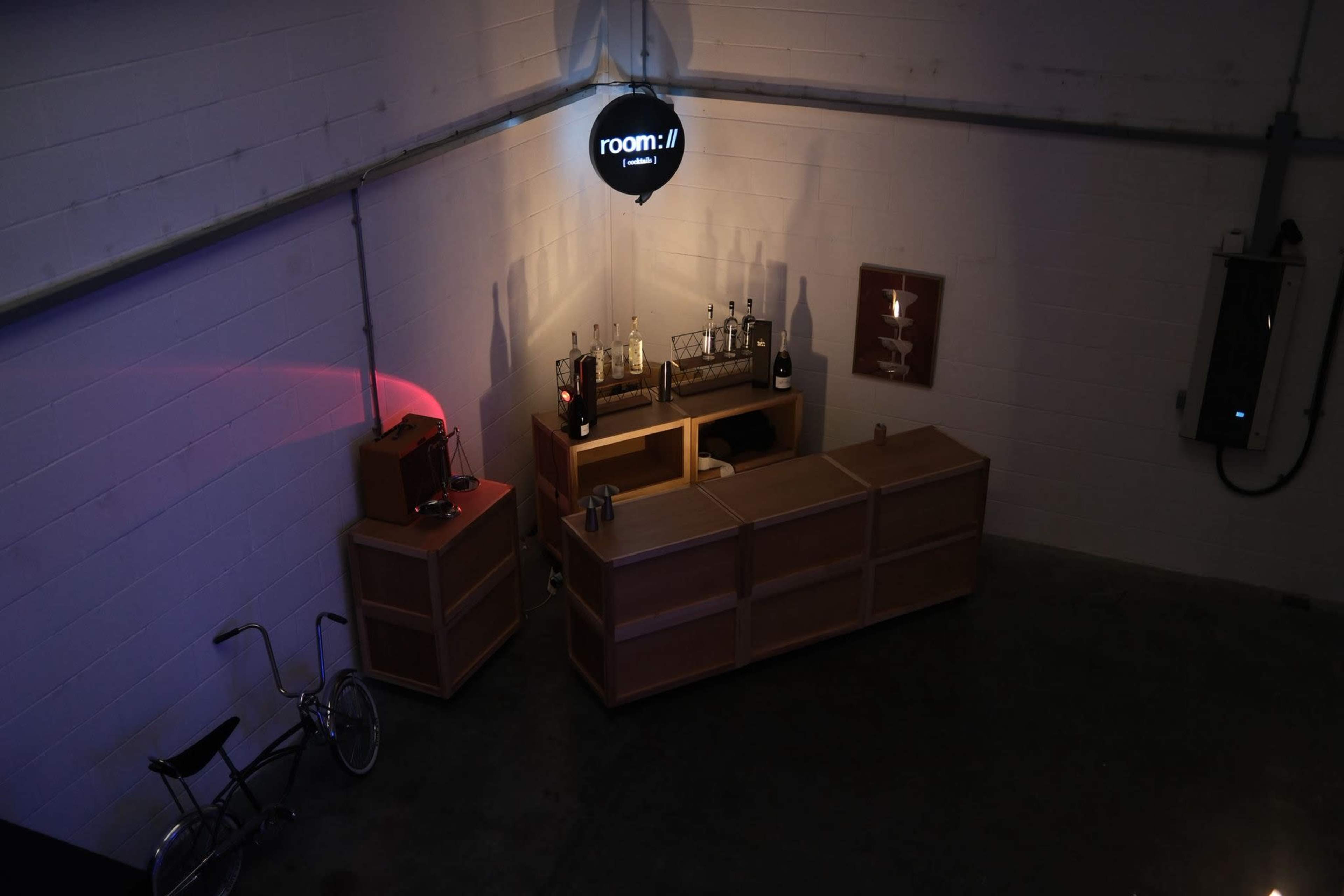 The image shows a dimly lit corner of a room featuring a bar with bottles, a wooden counter, and a bicycle placed nearby.