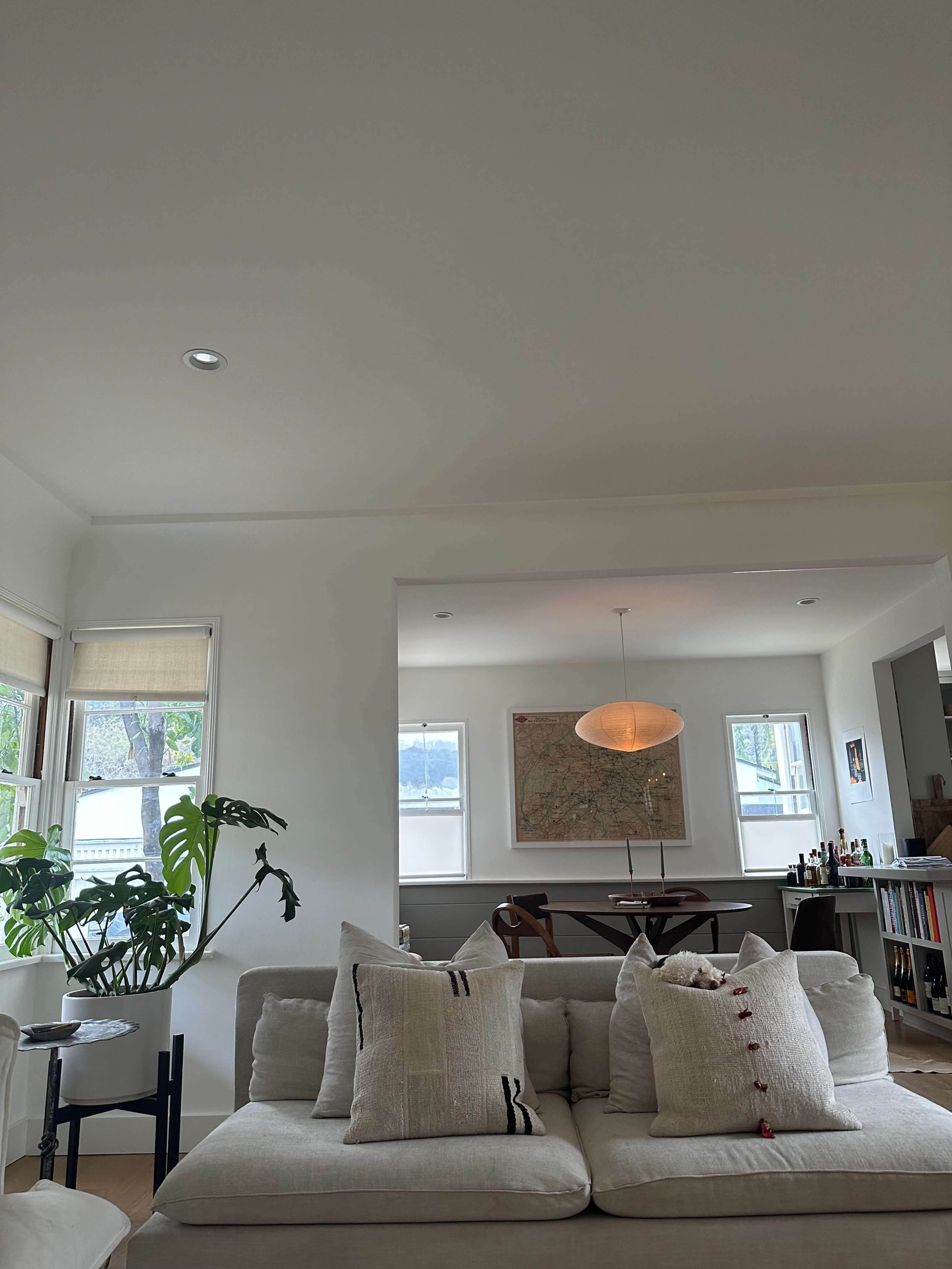 A living room with a light-colored sofa adorned with decorative pillows, a dining area in the background, and large windows allowing natural light to enter.