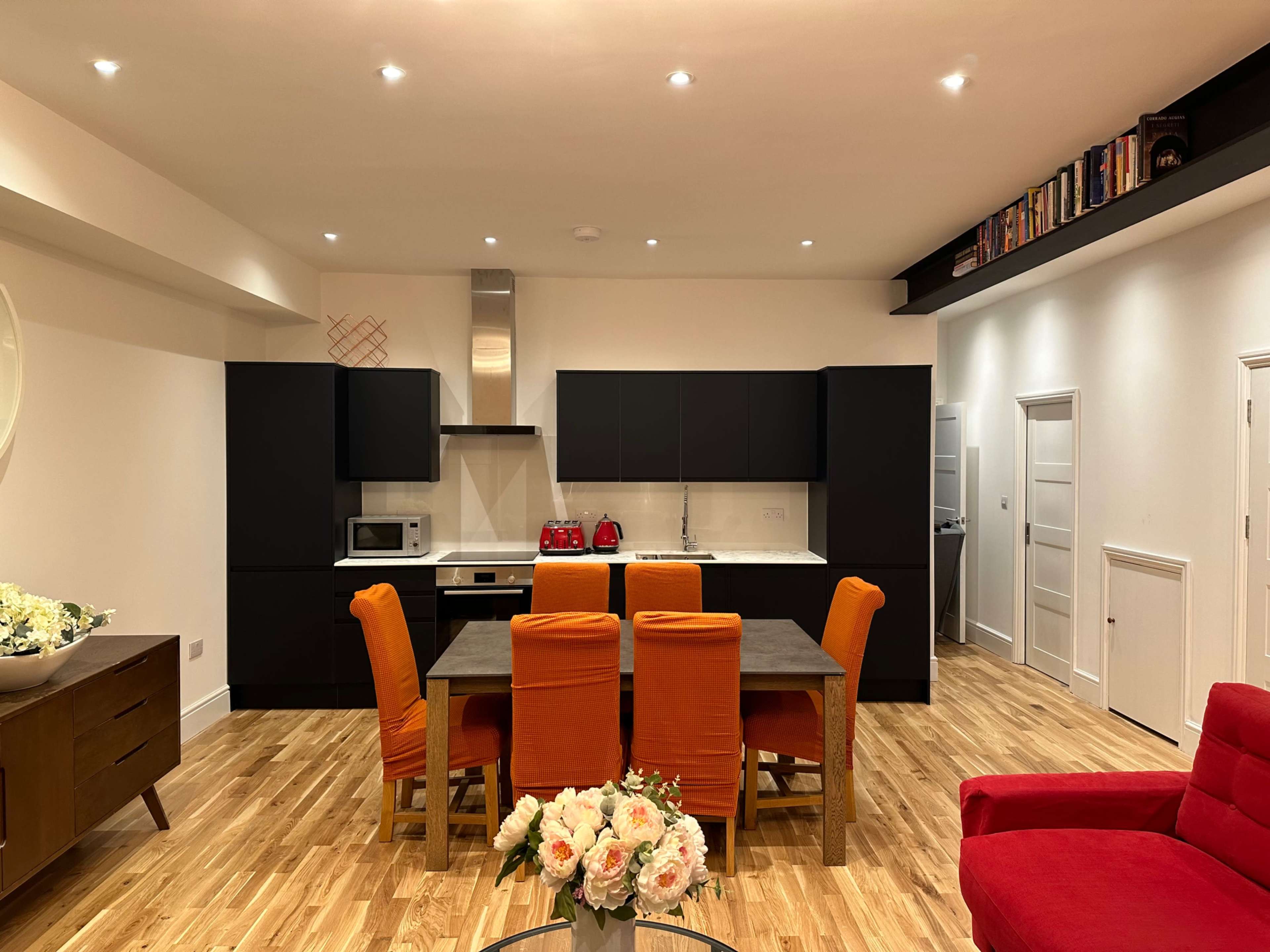 A modern kitchen and dining area features black cabinetry, a wooden table surrounded by orange chairs, and a red sofa in a well-lit space with hardwood flooring.
