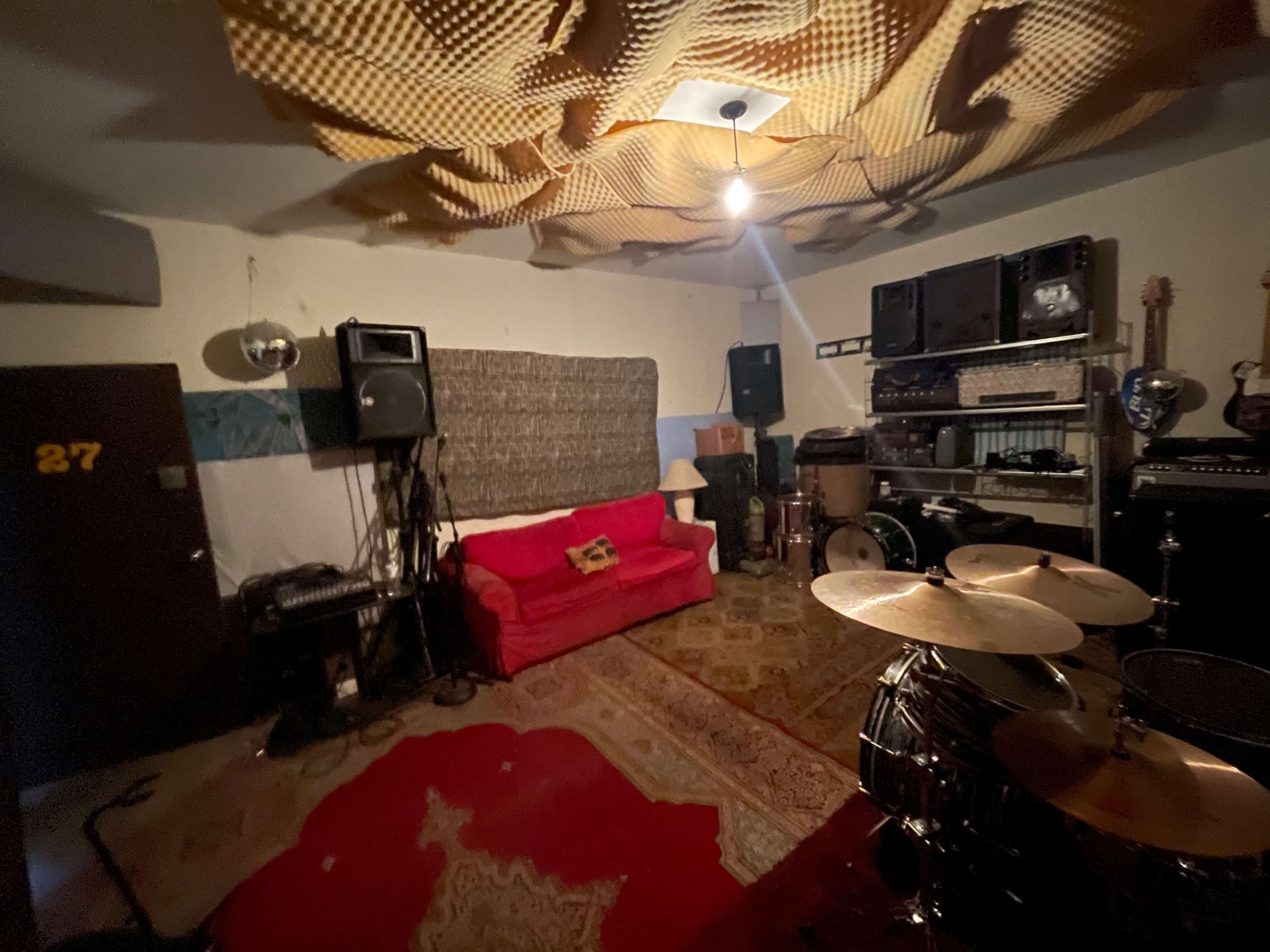 The image shows a musical rehearsal space featuring a red couch, various audio equipment, a drum set, and a carpeted floor.