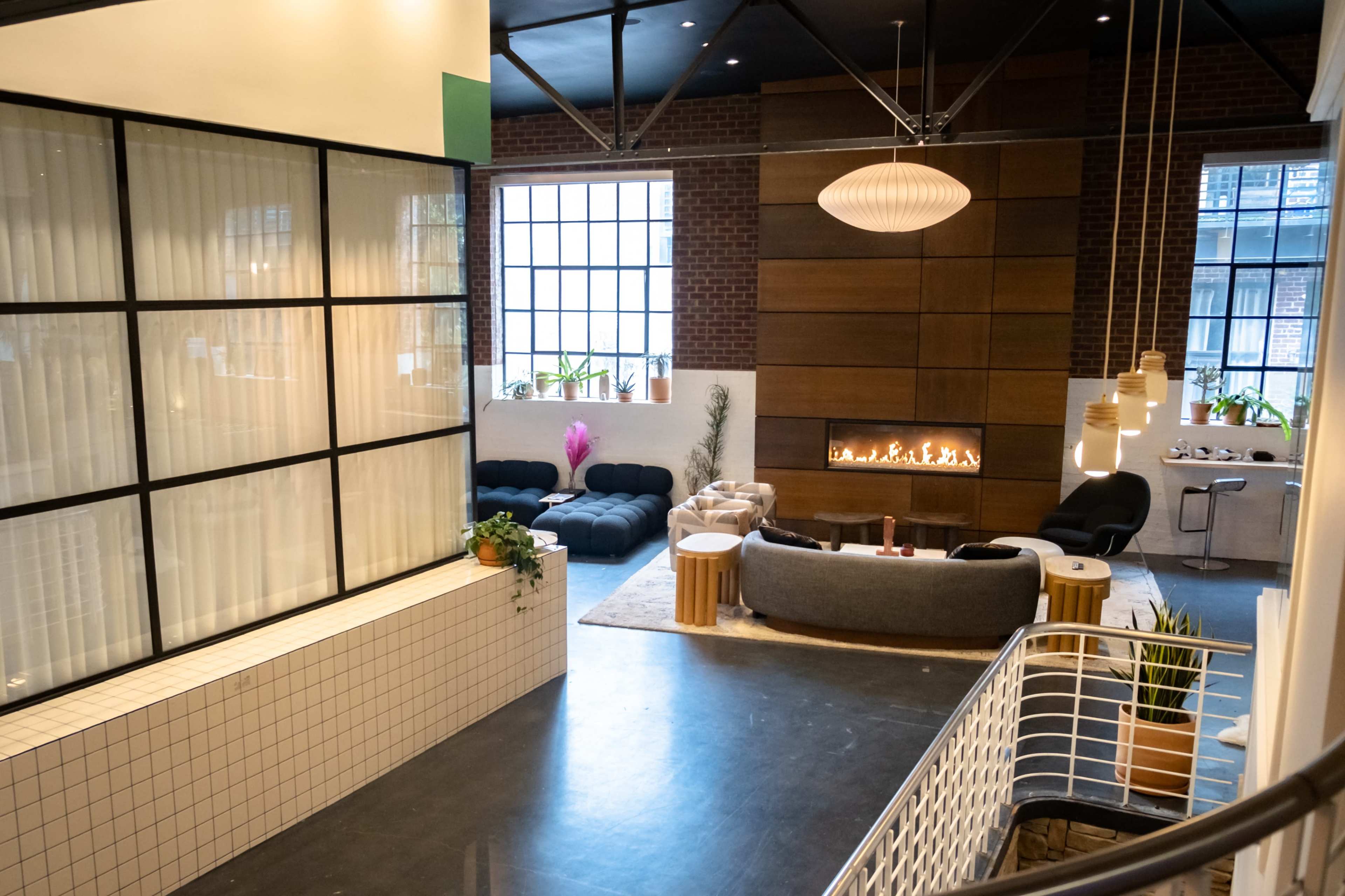 The image shows a modern, open interior space featuring a seating area with a fireplace, large windows, and a mixture of decorative plants and furnishings.