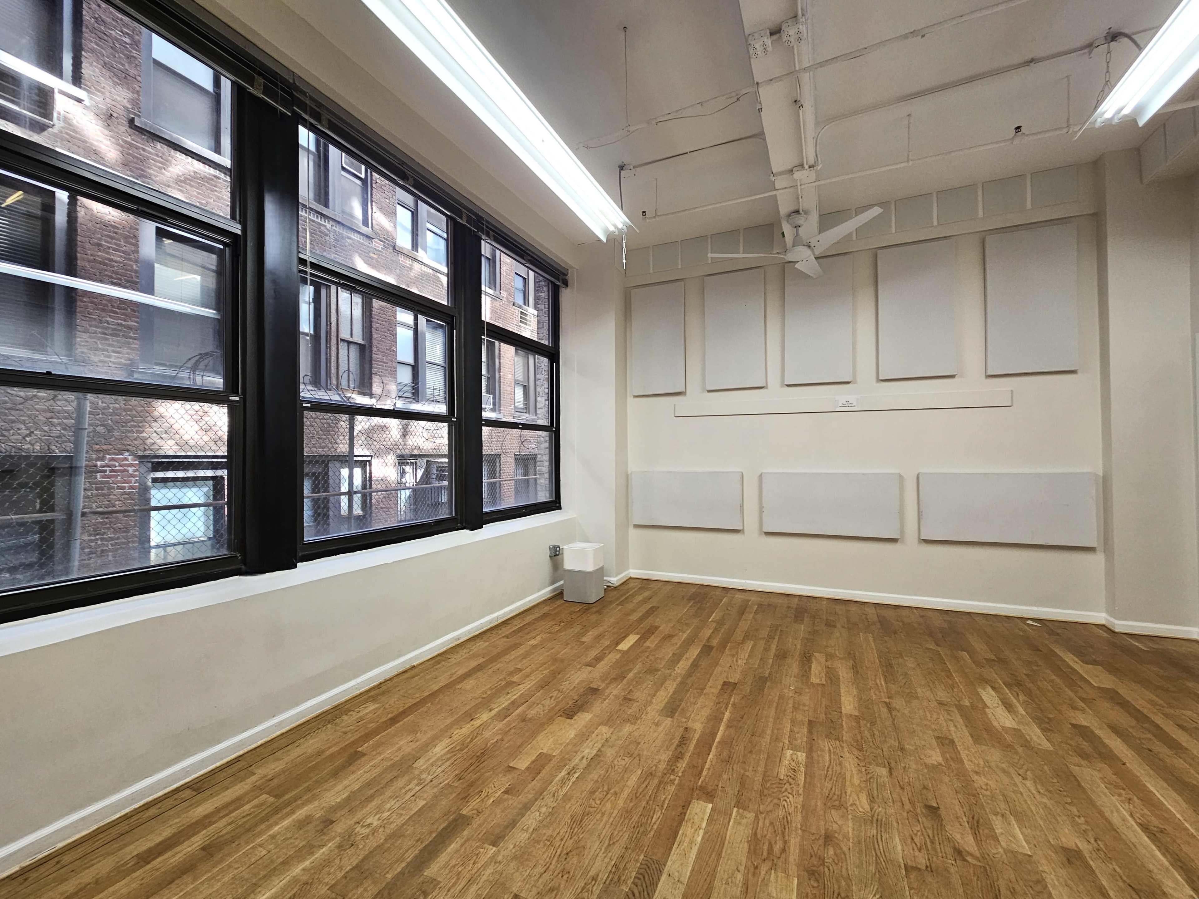 The image shows a spacious indoor area with large windows, wooden flooring, and blank wall panels.